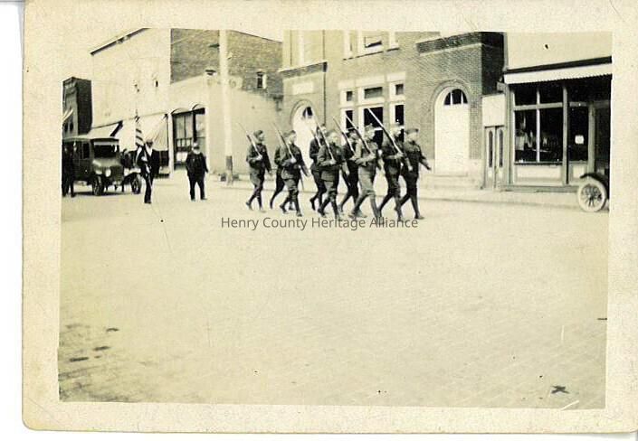 The vintage photograph portrays a group of soldiers marching in formation down East Street in Liberty Center, OH. They carry rifles on their shoulders and are led by a man holding an American flag, indicating this could be a parade or ceremonial procession. The soldiers are dressed in WWI uniforms. Surrounding them, early automobiles are parked along the sidewalk, and brick buildings line the street, evoking a historical period. A solitary pedestrian observes the march from a distance, adding to the quiet and solemn atmosphere. The scene captures a moment frozen in time, perhaps Memorial Day in 1920, reflecting a sense of discipline, patriotism, and community in the context of the era depicted.