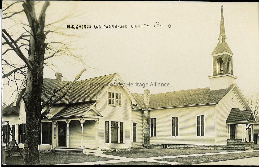 The image depicts a charming vintage photograph of Liberty Center's Methodist Episcopal church and parsonage. The church features classic architectural elements, including a prominent steeple with a bell tower, typical of many American small-town churches. The building is constructed in a simplistic, yet elegant style, characterized by its white clapboard siding and ornate detailing under the gables. Adjacent to the church is the parsonage, a residence for the church pastor, maintaining the same architectural style and color, suggesting a cohesive design throughout the property. A large, bare tree stands in the foreground to the left, providing a sense of scale and situating the buildings in a likely winter or early springtime setting. The image captures a moment in time and reflects the importance of such establishments in the historical fabric of small community life in rural America, showcasing the central role of religious and communal buildings during this era. The handwritten label at the top further personalizes the image, giving it an archival feel