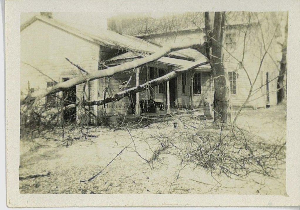 The image shows an old, weathered house with a large tree branch that has fallen and landed on the roof and front porch, indicating recent storm damage. The branches are sparse, with twigs extending in various directions, casting shadows on the ground, and the house's wooden siding and windows appear aged, adding an overall sense of vintage charm. Despite the potentially destructive scene, the photograph seems to capture a serene moment, with no visible humans or animals, just the quiet aftermath of nature’s force.