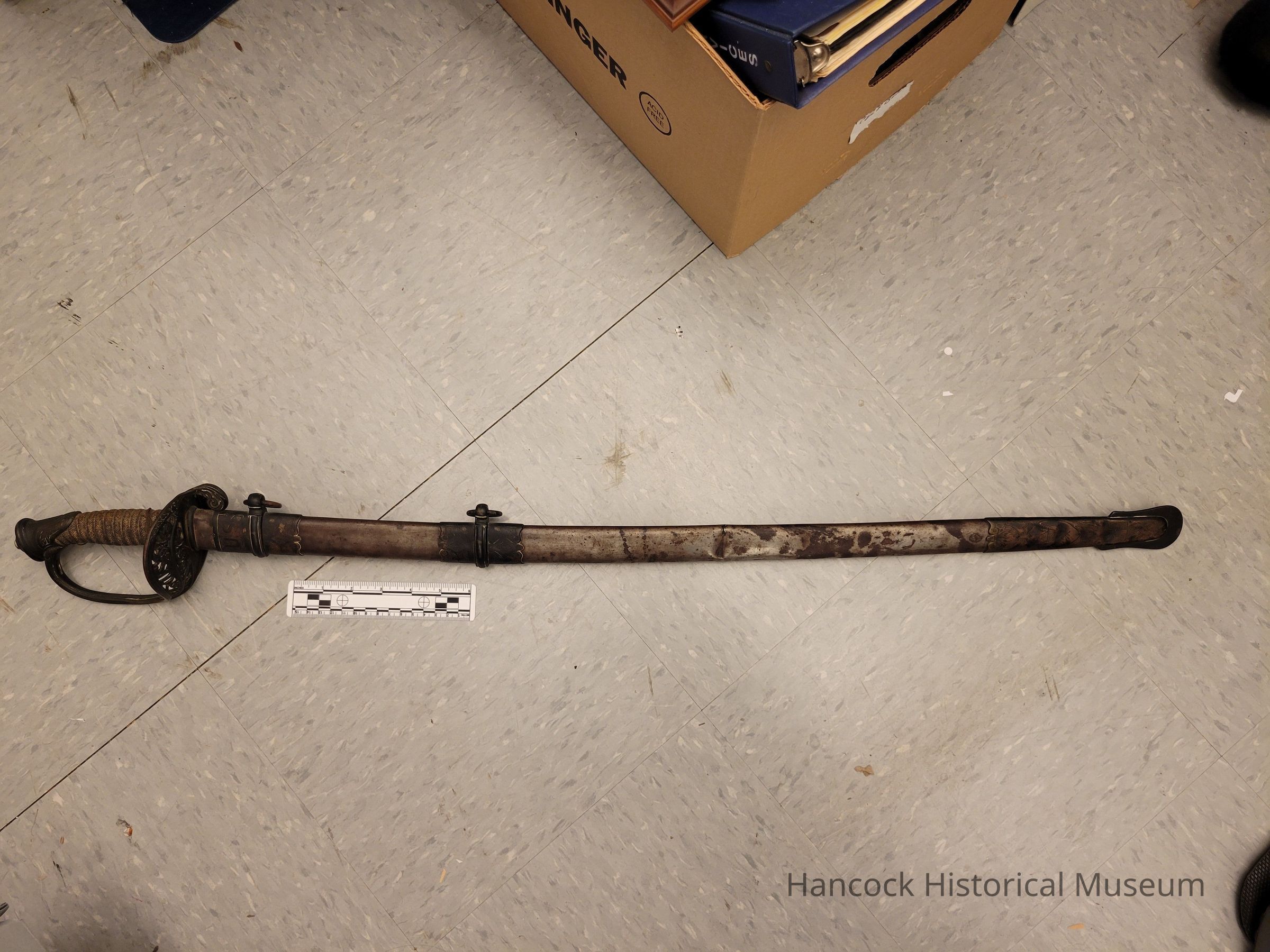 The image showcases a Civil War-era sword attributed to William Mungen, a colonel of the 57th Ohio Volunteer Infantry (OVI). The sword is encased in a weathered scabbard, exhibiting signs of age and historical usage, with a distinguishable patina along its length. The hilt features intricate guard designs, with a grip that appears to be wrapped or covered, suggesting practical use during its time. Positioned on a tiled floor, the sword's placement is aligned with a measuring scale to provide context for its size.