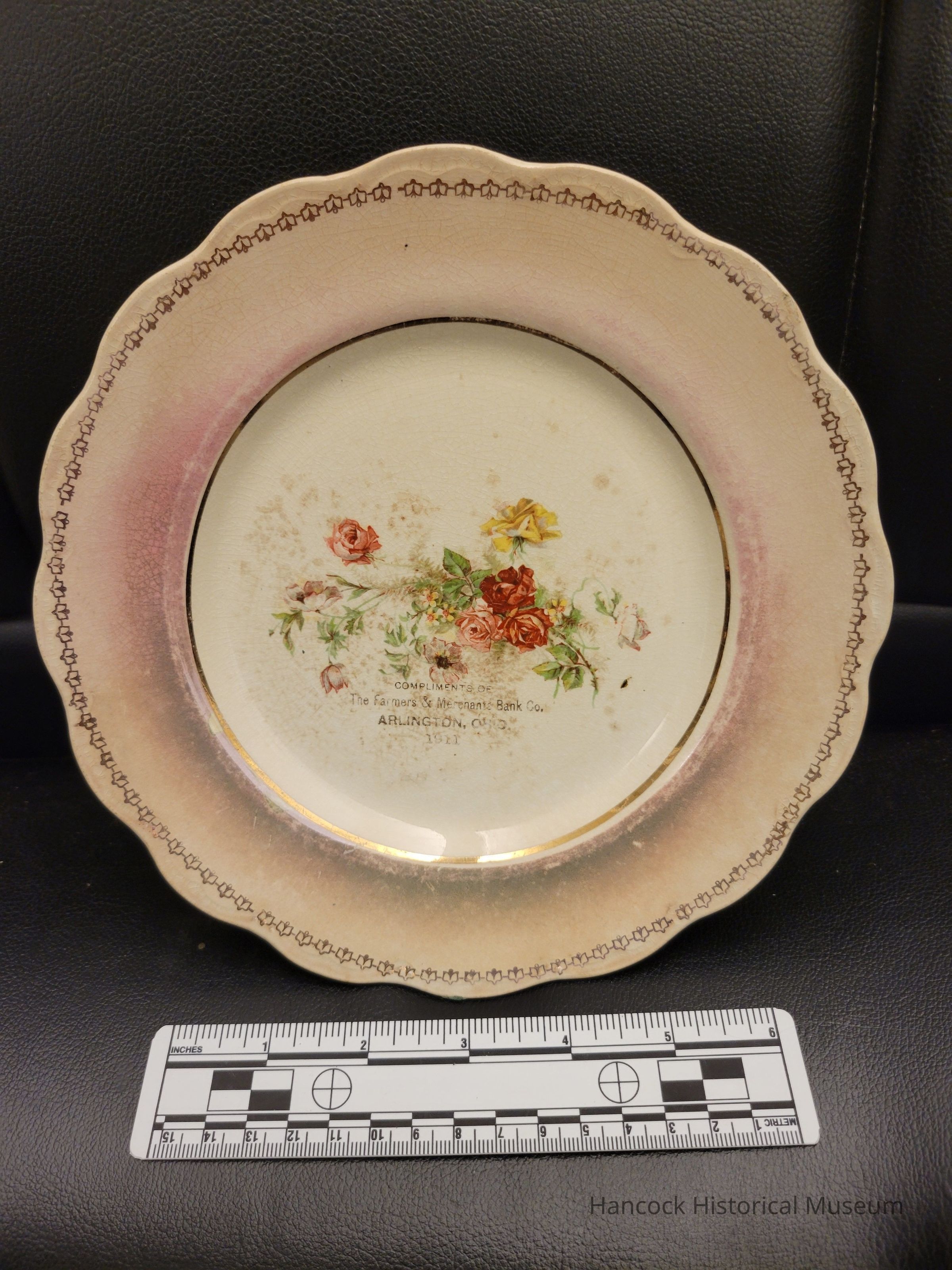 The image features an ornate decorative plate resting on a dark surface, which is embellished with an intricate floral pattern of pink, red, and yellow flowers at its center, exuding a vintage charm. The plate's border, tinged with a subtle pink hue, is highlighted by a repeated pattern of small, interconnected geometric designs around its gently scalloped edges. A central inscription reads: "COMPLIMENTS OF The Farmers & Merchants Bank Co., ARLINGTON, O. 1921," suggesting it might have been a commemorative or promotional item from that time period. Positioned beneath the plate, a detailed ruler with both metric and imperial measurements is visible, likely provided for scale or to authenticate the image.