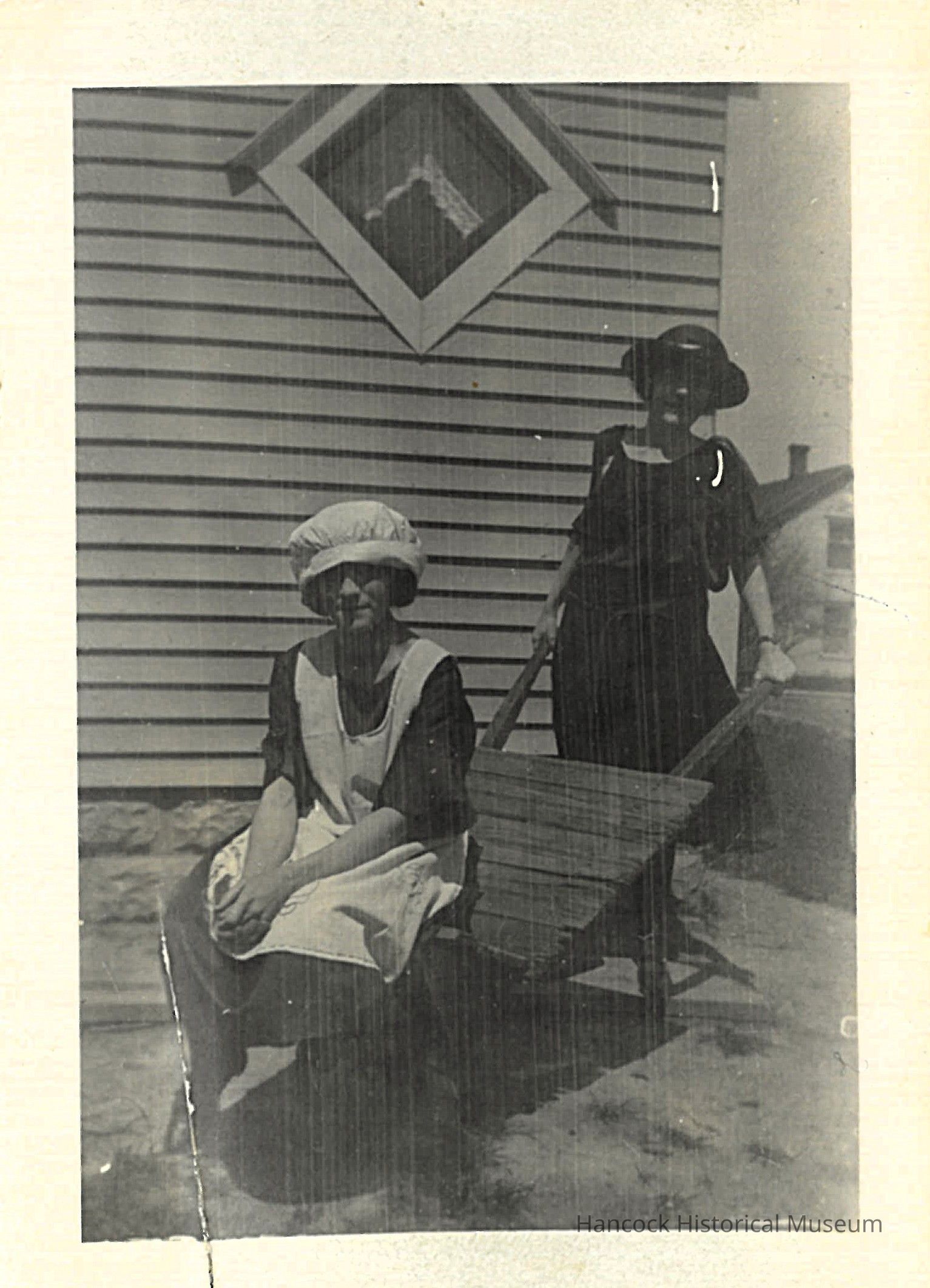 The black and white photograph presents two women dressed in early 20th-century attire, standing in front of a building with clapboard siding and a diamond-shaped window adorned with curtains. The woman in the foreground is seated on what appears to be a wooden wheelbarrow, wearing a dark dress with a white apron and a bonnet, gazing directly at the camera. Behind her, another woman stands, dressed in a dark outfit with a wide-brimmed hat, holding the handles of the wheelbarrow, and casting a shadow on the wall.