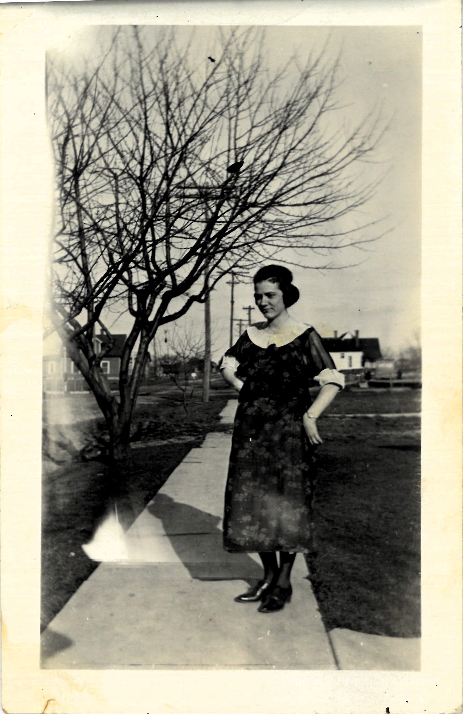 In a black-and-white photograph, a woman stands poised on a paved walkway with hands on her hips, wearing a dark floral dress and boots, exuding a confident expression as she is framed by a leafless tree to her left, with houses in the background and clear sky above, capturing a moment of early 20th-century life amidst a suburban setting.
