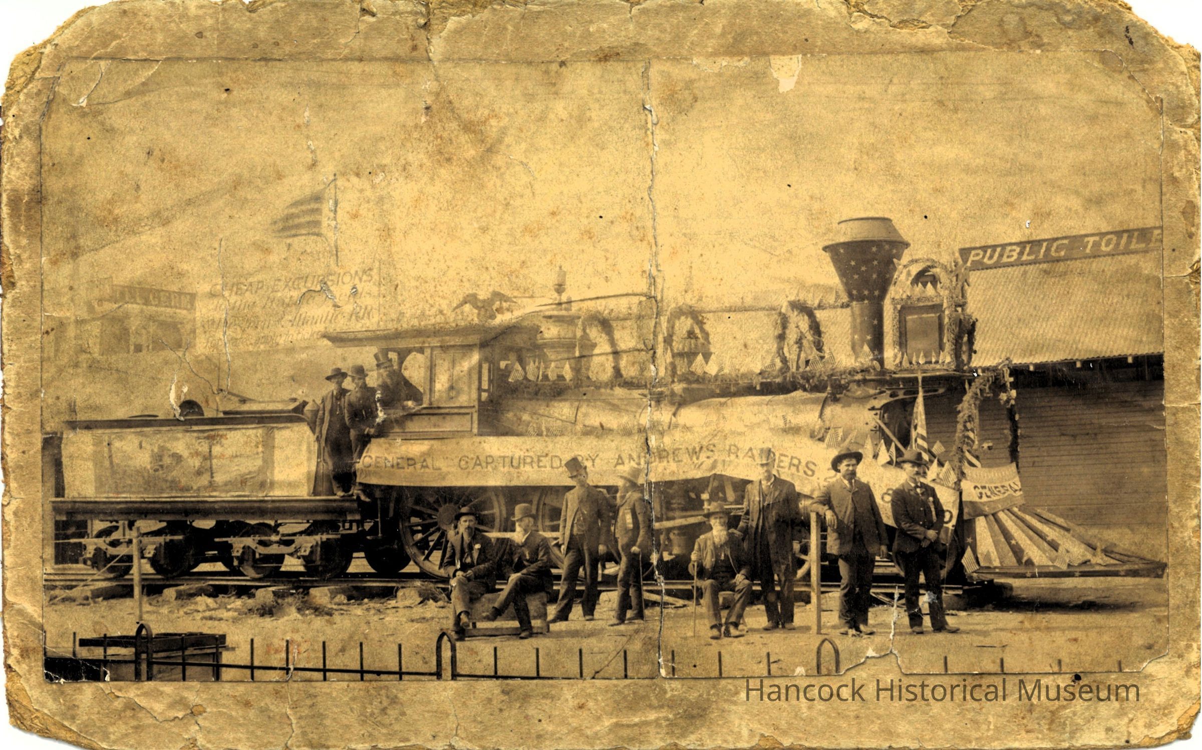 The image shows the renowned locomotive "General," famously associated with Andrews' Raiders during the Civil War, positioned on railway tracks. A group of men, dressed in period attire, stands or sits near the locomotive, some leaning against it. The engine is adorned with decorations, emphasizing its historical significance. A banner prominently displayed on the side of the locomotive reads "General, Captured by Andrews' Raiders." In the background, a building and the partially visible sign suggest a public space, hinting at the importance of the event.