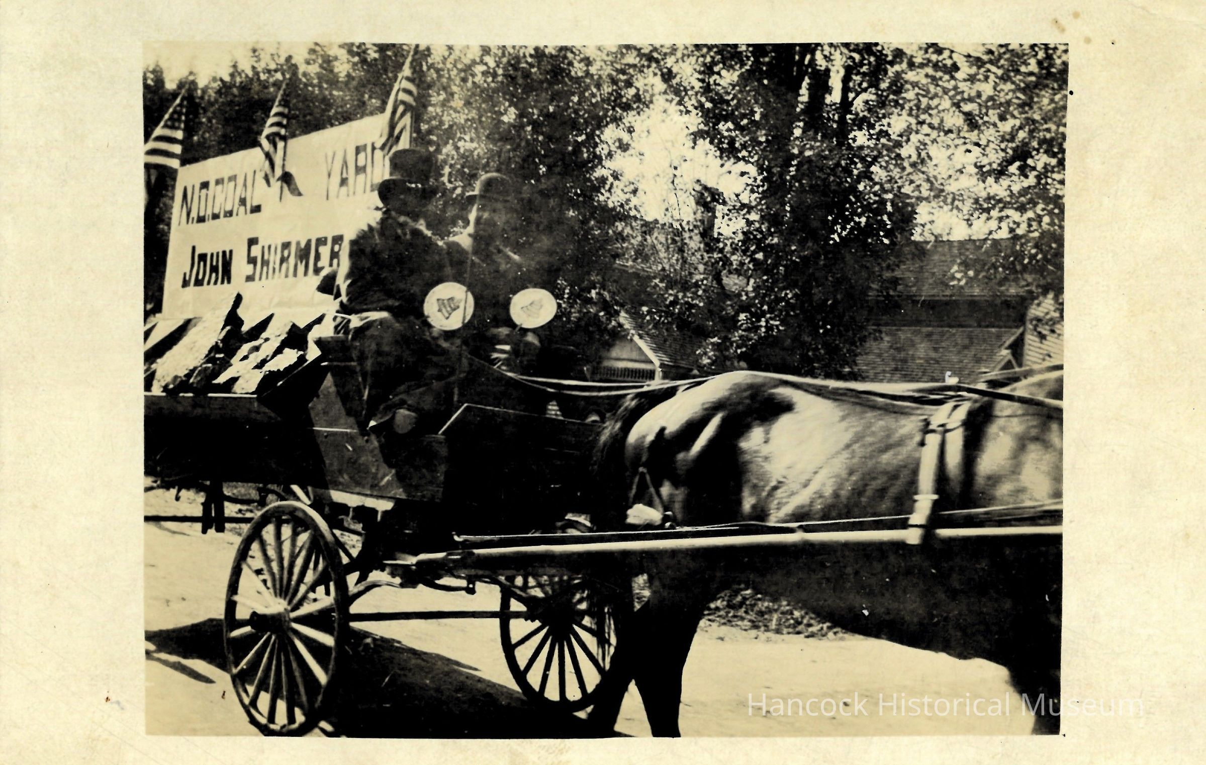 The image depicts a vintage black-and-white scene featuring a horse-drawn carriage carrying two individuals dressed in period attire, with one person wearing a top hat and the other in a bowler hat. The carriage is part of a parade or procession, as indicated by multiple American flags displayed in the background. Behind the carriage, a large sign can be seen, which reads “NO COAL YARD” and includes the name “JOHN SHIRMER.” The road appears unpaved, with trees lining the sides, contributing to the rustic and historical ambiance of the setting. The sun casts shadows, suggesting a clear day, and the carriage’s wheels are prominently visible in the foreground.
