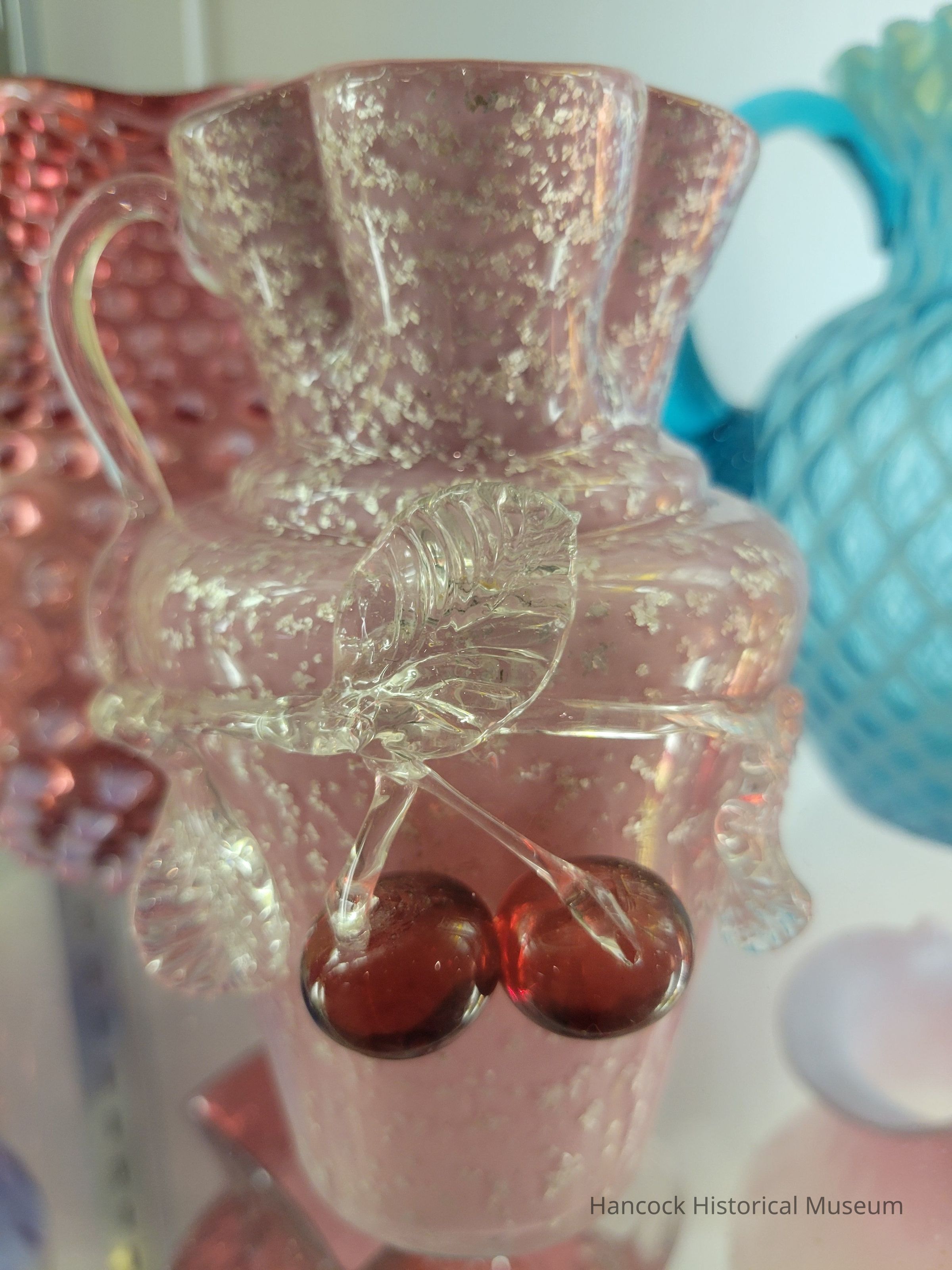 The image showcases a delicate glass vase with a pinkish hue and textured surface, adorned with intricate details like clear glass leaves and two shiny red cherries, set against a varied background featuring other colorful glass pieces that provide an artistic and vintage ambiance to the overall composition.