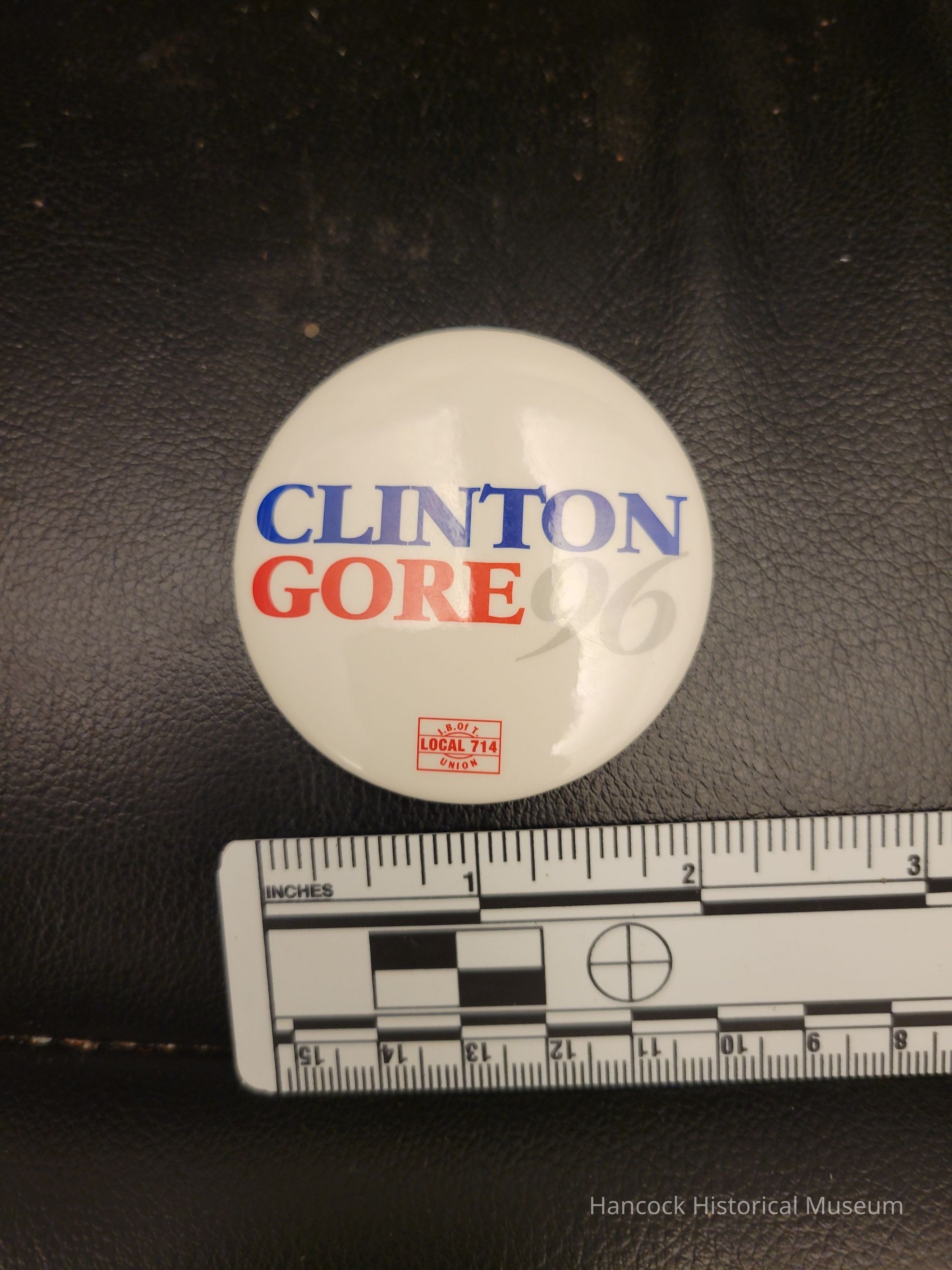 The image displays a campaign button from the 1996 U.S. presidential election, featuring the names "Clinton" and "Gore" in bold blue and red text on a white background, with "96" in gray; a small emblem reading "U.B.O.T Local 714 Union" is printed beneath, and a white ruler marked in inches is placed below the button on a black leather surface to provide scale.