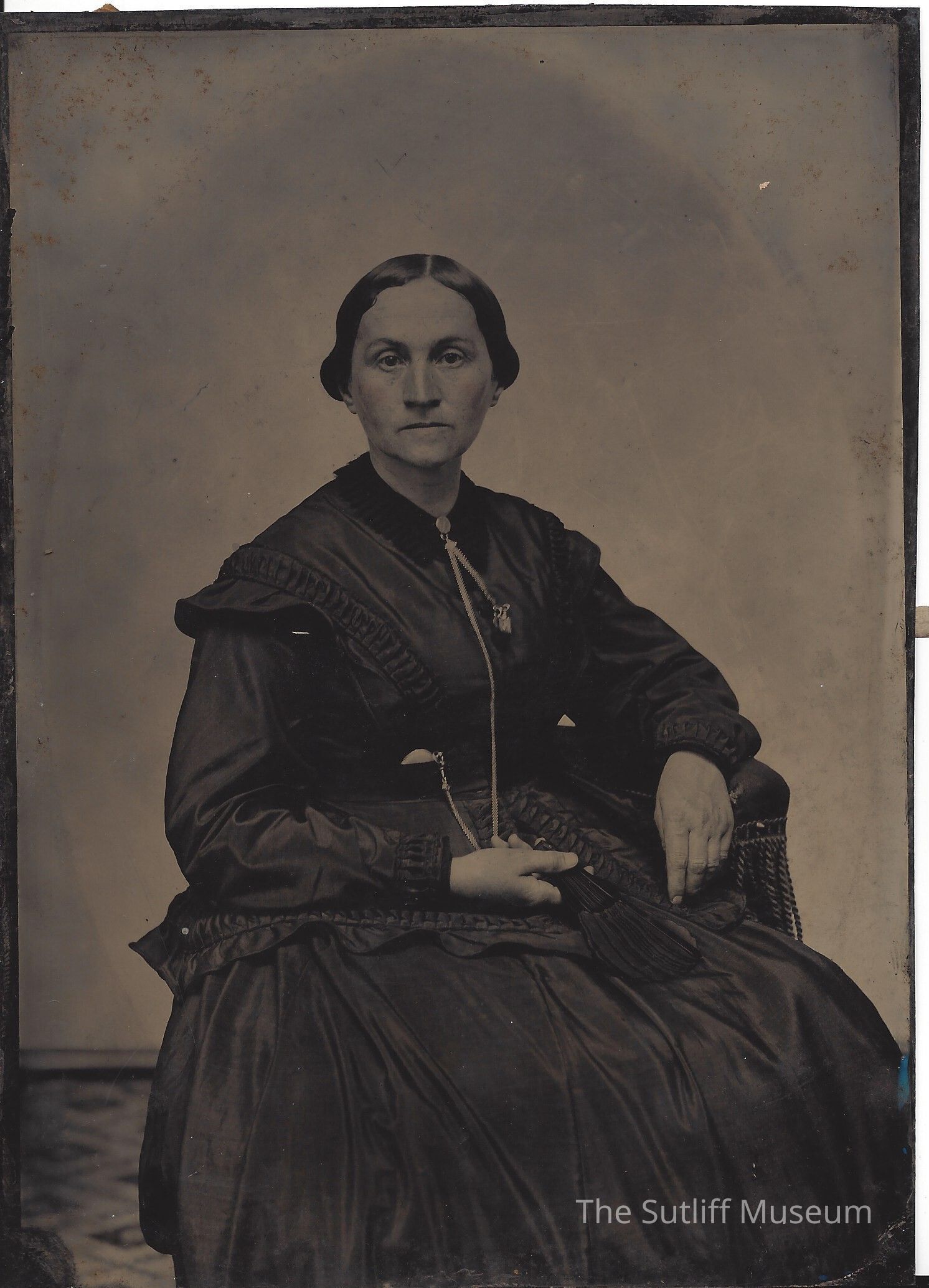 1971.31.182 front: The photograph features a woman seated in an ornate wicker chair, dressed in a dark, voluminous 19th-century gown with detailed ruching and pleating. She wears somber expression with her hands positioned gently in her lap, one holding a fan. Her attire is complemented by a distinct, long necklace that falls to her waist, decorating her otherwise plain dress. This sepia-toned image, showing some signs of age and wear, captures an essence of bygone elegance.