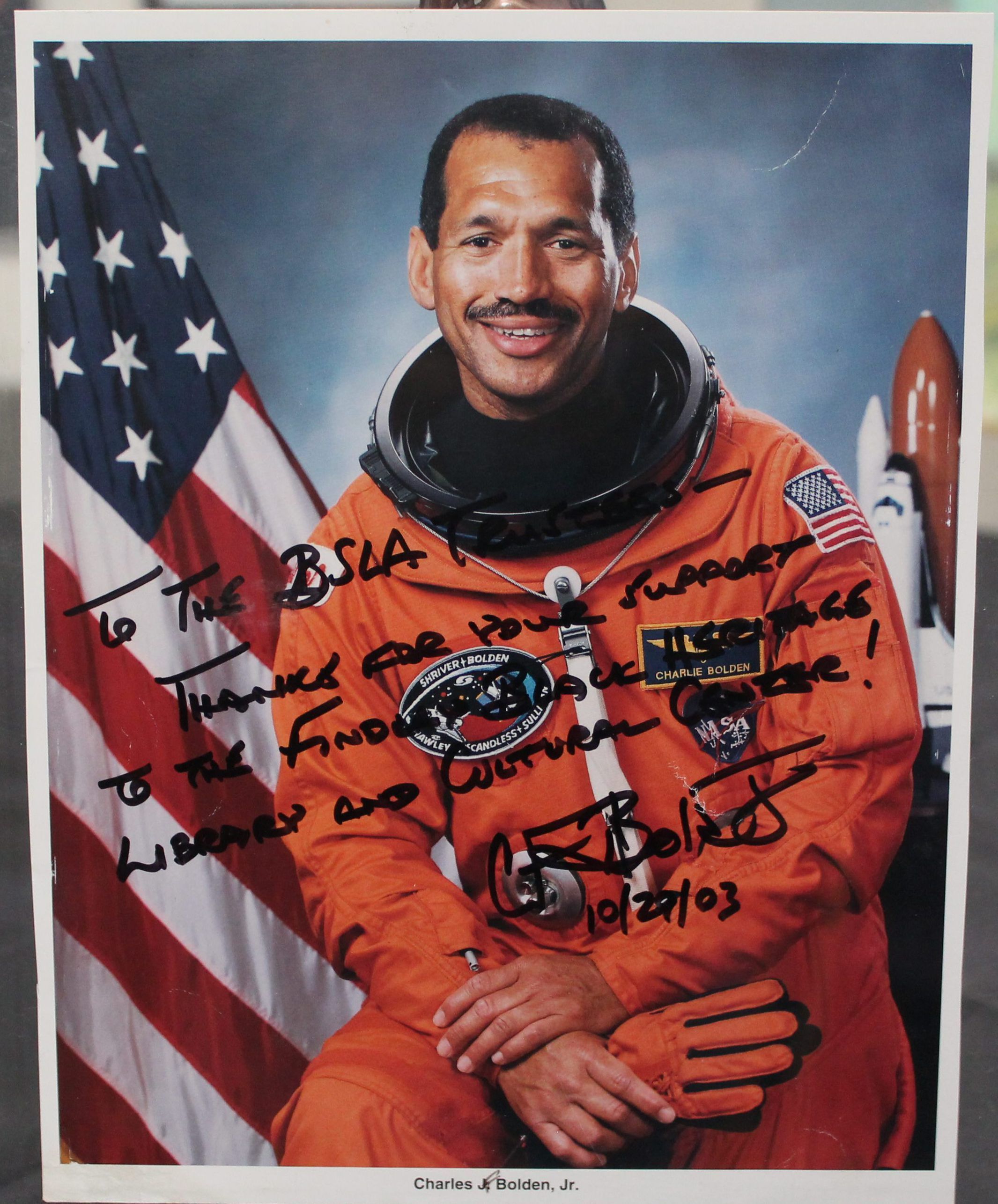 Front of image: The image depicts Charles F. Bolden, Jr. in his orange astronaut space suit. In the background, the American flag is on the left of the image, and his spaceship on the right. At the bottom of the picture was written: "Charles J. Bolden, Jr." the middle initial "J" has been replaced with an handwritten "F".
The inscription reads:
To The BSLA -------(?),
Thanks for your support
to the Findlay Black Heritage Library and Cultural Center!
CFBolden
10/20(?)/03