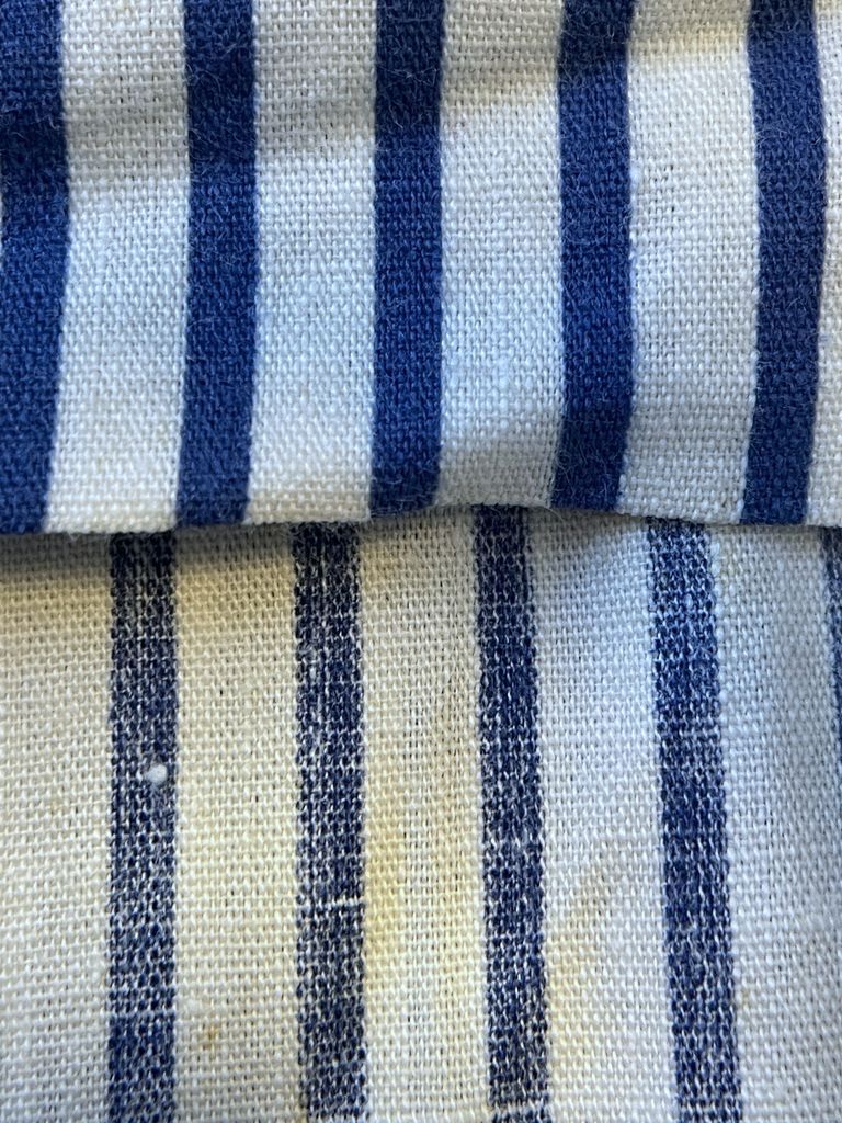 The face and reverse sides of the fabric shown side by side — The top half of the image shows the face of a plain-woven fabric. There are solid-colored, equidistant stripes in a navy color running vertically along the fabric. The bottom half shows the reverse side. The stripes are slightly patchy, and several slubs are clearly visible.