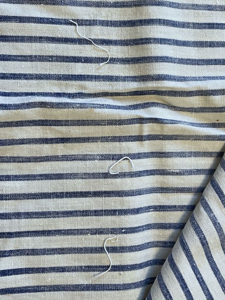 The reverse side of the fabric, showing unpicked threads — Image showing the reverse side of a plain-woven fabric. Navy blue stripes run horizontally along the fabric. Three small pieces of thread are intertwined with the piece, running perpendicular to the stripes.