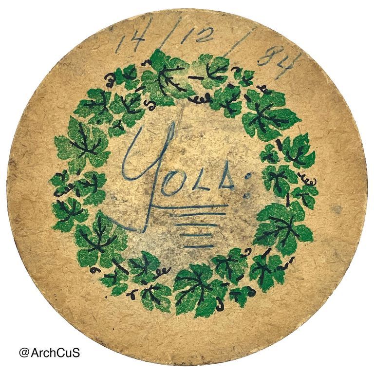          Circular coaster made of brown fiber with a circular design of an ivy wreath in the middle. A written note in the center spells 