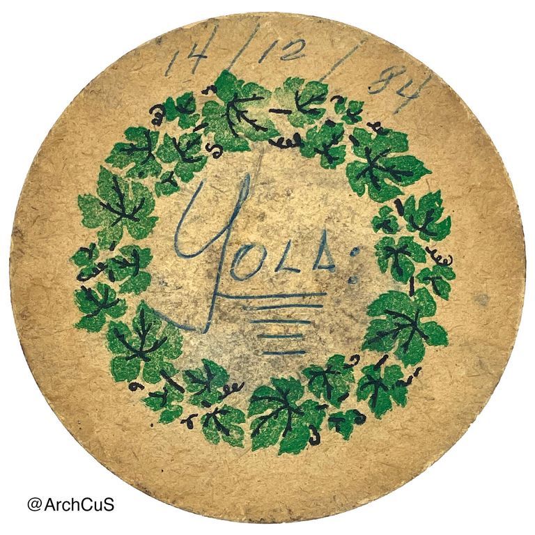 Circular coaster made of brown fiber with a circular design of an ivy wreath in the middle. A written note in the center spells "Yola," written in blue pen ink.