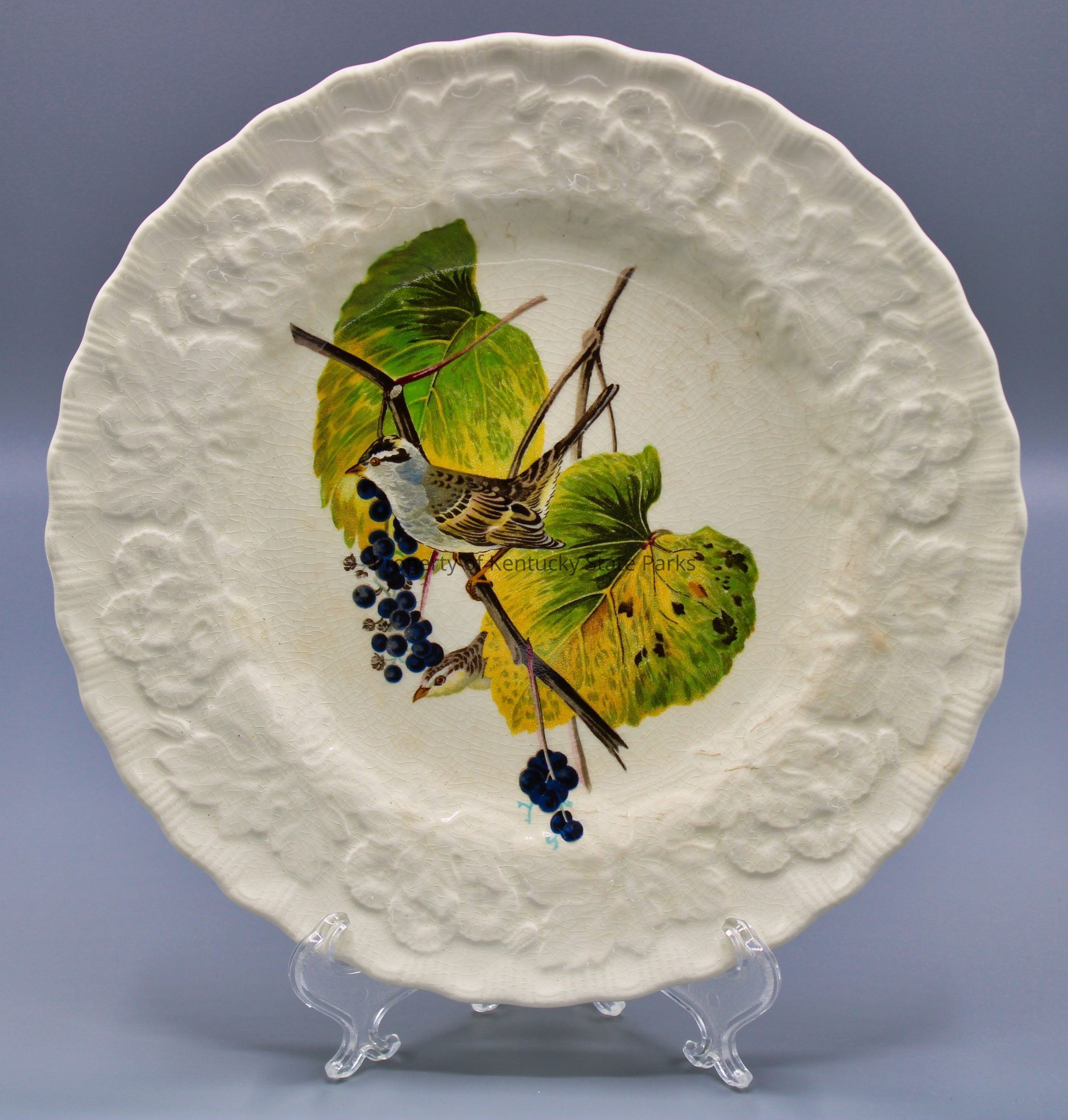 Luncheon plate with reproduction of John James Audubon's "White-crowned Sparrow": A white china plate with a raised pattern of flowers, scalloped edges, and a color image of a bird perched on a branch with blue berries and greenish-yellow leaves. The bird faces to the viewer's left. Its feathers are colored brown, gray, white, and black. Photograph date: March 30, 2023 / Photographer: Heidi Taylor-Caudill