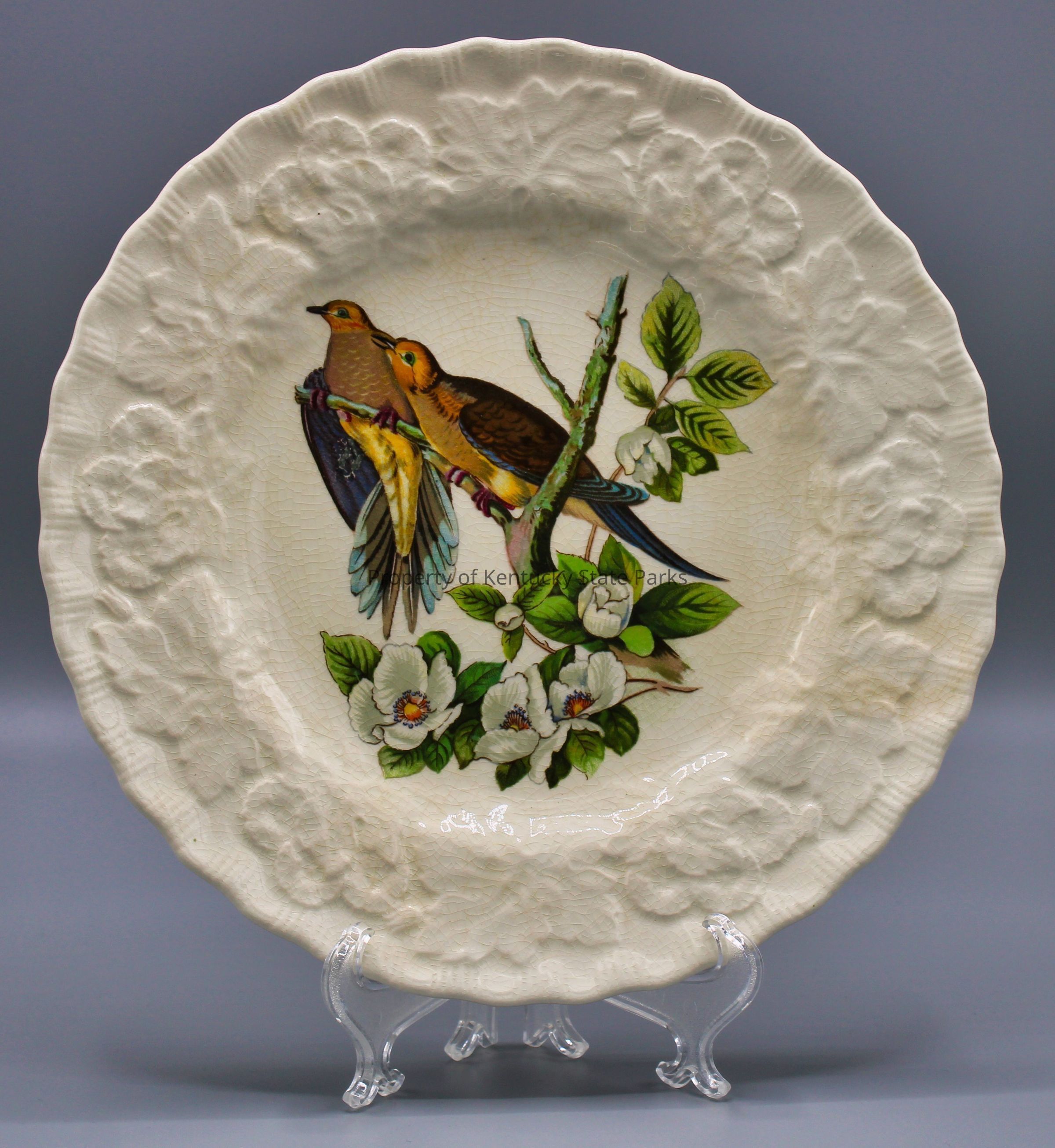 Luncheon plate with reproduction of John James Audubon's "Carolina Turtle Dove," also known as the "Carolina Pigeon": A white china plate with a raised pattern of flowers, scalloped edges, and a color image of two birds perched on a branch of white flowers and green leaves. The birds face to the viewer's left. Their feathers are colored brown, yellow, blue, purple, and orange. Photograph date: March 30, 2023 / Photographer: Heidi Taylor-Caudill