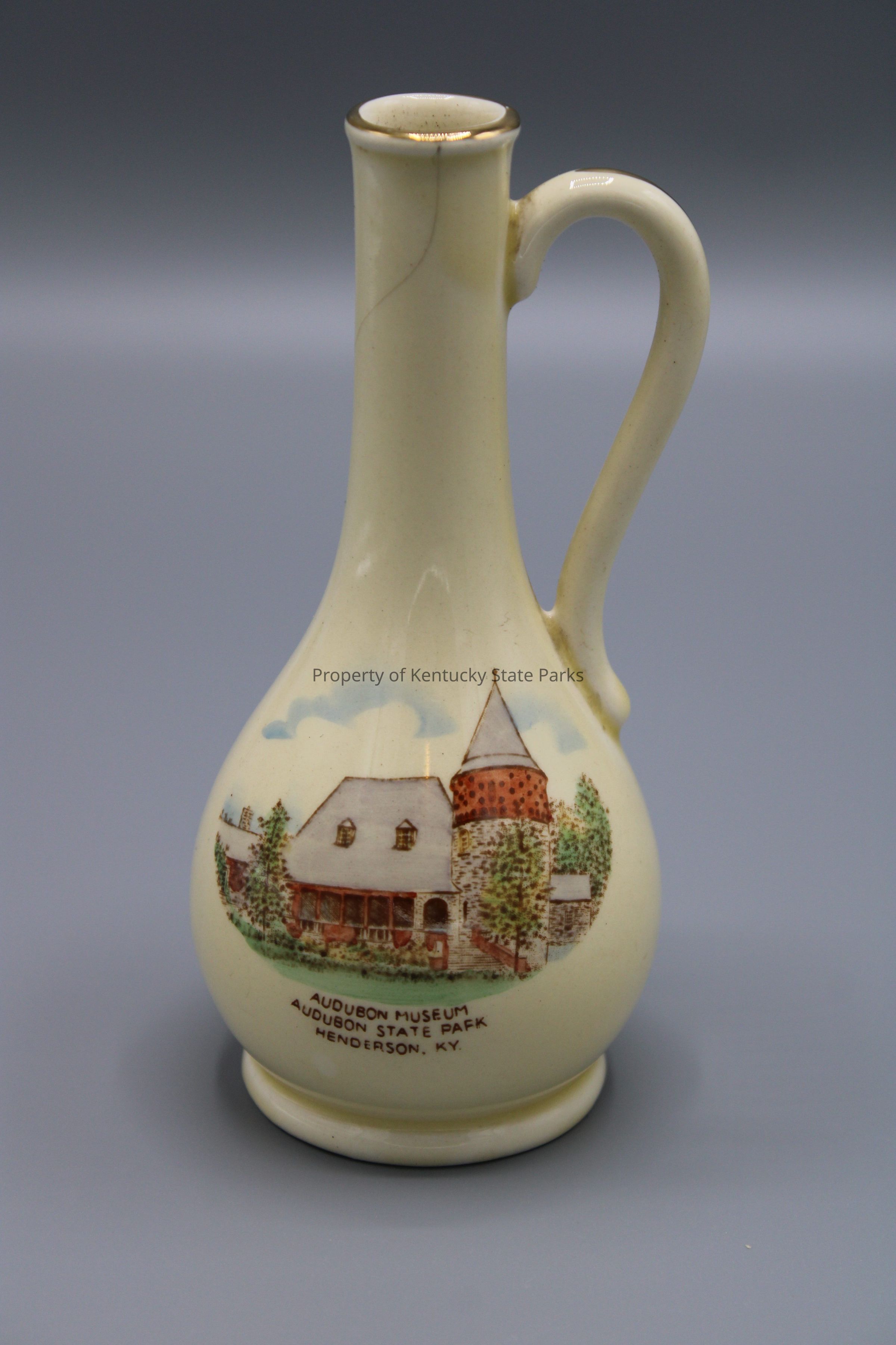 The image depicts a cream-colored ceramic pitcher with a slender, elegant form and a curving handle. On its surface, there's a detailed, picturesque illustration featuring a scenic depiction of the Audubon Museum located at Audubon State Park in Henderson, Kentucky. The artwork showcases the museum building with its distinctive architecture, partially surrounded by lush green trees, under a soft blue sky with fluffy clouds. The illustration is rich in detail and color, adding a charming and vintage look to the piece. The overall aesthetic of the pitcher exudes a sense of historical and rustic beauty, making it not only a functional item but also a decorative piece that captures a specific locale's essence.