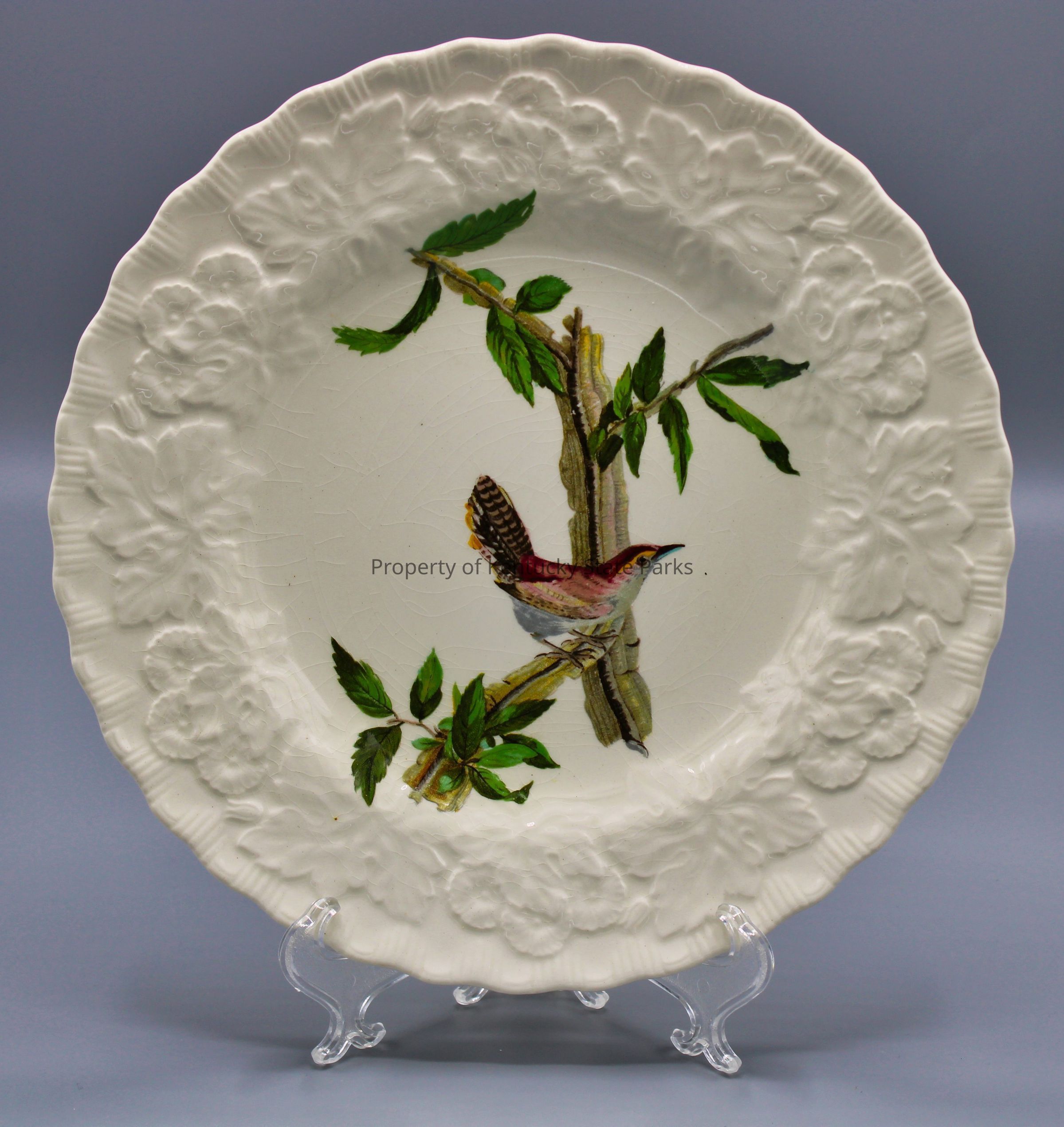 Luncheon plate with reproduction of John James Audubon's "Bewick's Wren": A white china plate with a raised pattern of flowers, scalloped edges, and a color image of one bird perched on a branch with green leaves. The bird faces to the viewer's right. Its feathers are colored red, pink, yellow, gray, black, and brown. Photograph date: March 30, 2023 / Photographer: Heidi Taylor-Caudill