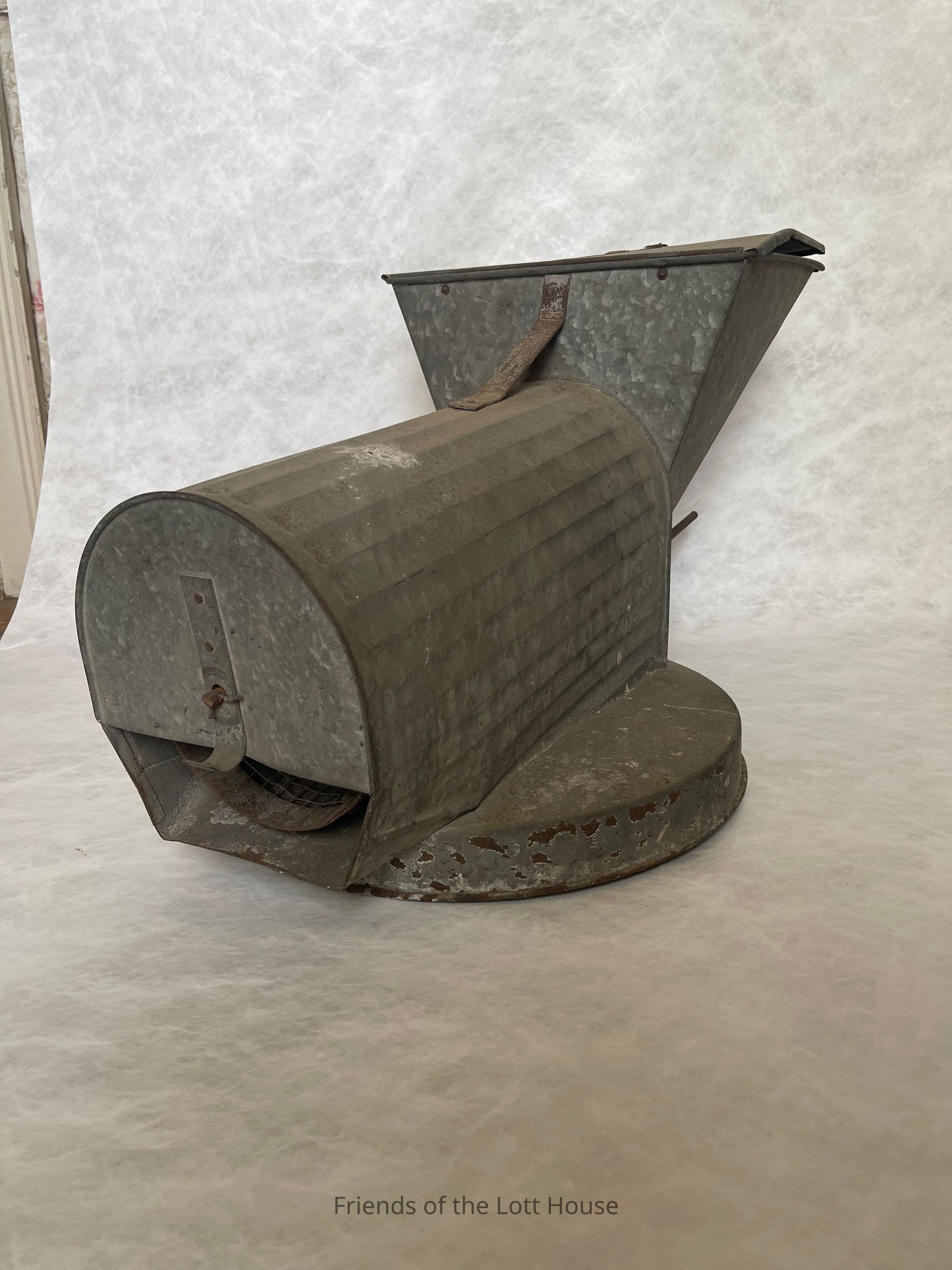 A feed grinder is a machine used in agriculture to grind or crush feed materials such as grains, seeds, and other ingredients used to make animal feed. This process helps to break down the feed into smaller particles, making it easier for livestock to digest and absorb the nutrients
People have used feed grinders for centuries, with the earliest examples dating back to the use of mortars and pestles. Feed grinders were particularly important during the 19th and early 20th centuries. Before the widespread use of grinders like this, farmers relied on more labor-intensive methods of preparing feed for livestock, often grinding by hand or using makeshift tools. The grinder revolutionized farm animals' feeding, increasing agricultural efficiency and productivity.
This particular grinder from our collection dates back to the early-to-mid 20th century. Grains, corn, and other feed materials were put into the machine, while the hand crank turned the internal mechanism that ground the feed into smaller pieces. The barrel and round base ensured the device was stable during operation, and the lip was a thoughtful design choice to prevent spillage by allowing the grinder to rest securely on a container.
This feed grinder was made by NESCO — National Engineering and Supply Company—and features the logo “Young Patriot Boy.” The company was recognized for producing high-quality, durable agricultural equipment used by farmers across the United States from the 1860s to the 1950s. Two German immigrant brothers, Frederick G. Niedringhaus (1834-1922) and William F. Niedringhaus (1835-1908), founded the company; they were trained tinsmiths. The company was integral to the founding of Granite City, Illinois, as an industrial hub for iron and steel production. Innovations like enameling tin and political influence, particularly through protective tariffs, fueled NESCO's growth.
The company moved to Granite City in the 1890s, where it built extensive mills and helped develop the city. NESCO's success also supported local steel companies, making Granite City a major industrial center.