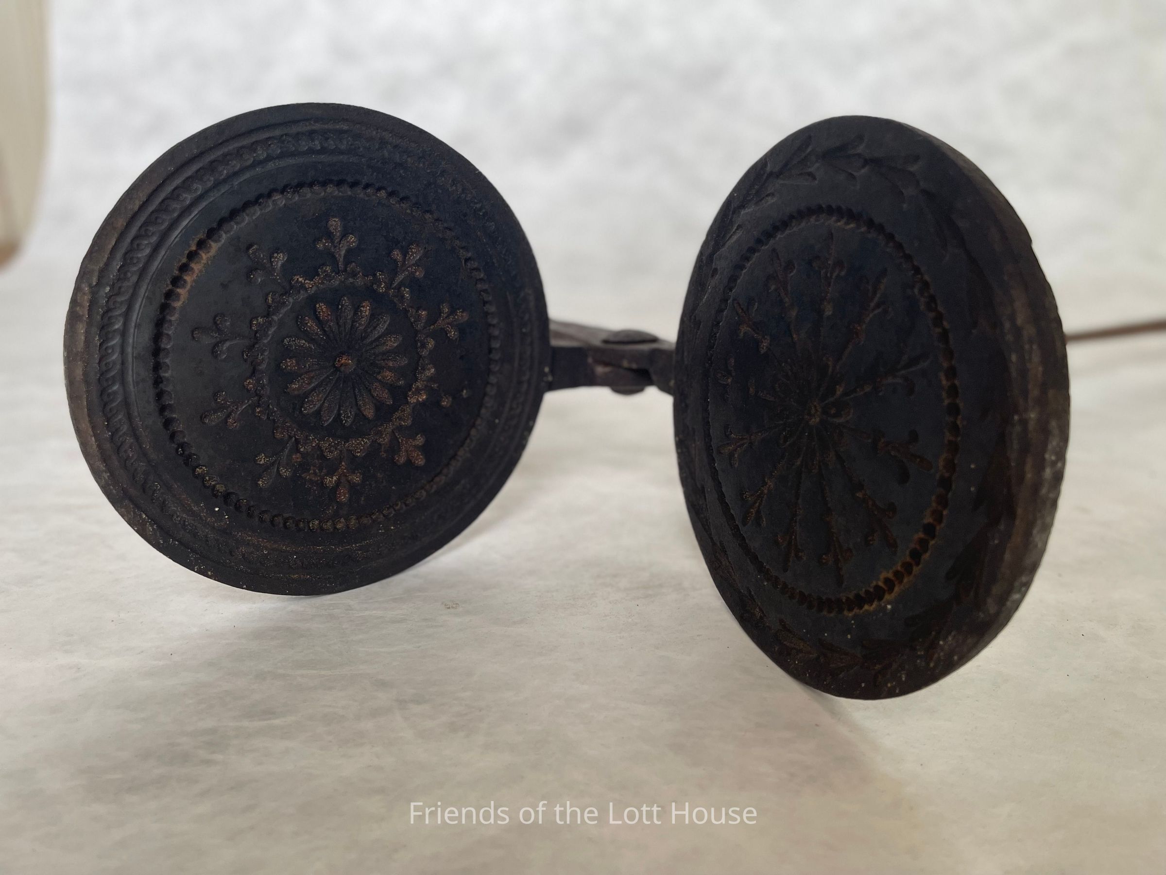 The plates of waffle irons were typically made of cast iron, a material common in kitchen tools of the time due to its durability and heat retention. The handles of waffle irons were long and typically made of wood or metal. The long handles helped the cook hold the hot iron over the fire without burning themselves. They often had decorative patterns etched into the metal, such as flowers, grids, or geometric shapes. This one from the Stuart and Annette Mont Collection, of objects from the Wyckoff-Bennett-Mont House, features two intricate patterns on each side of the plate.
They would place the waffle iron over an open flame. After it was properly heated, the cook would grease the plates with butter or lard to prevent them from sticking, and then pour the batter into the iron. The iron was then clamped shut and held over the heat for a few minutes, carefully turning it to ensure that both sides cooked evenly. The result was a crisp, golden waffle with a pattern from the iron plates. Waffle-making was a labor-intensive process, requiring careful attention to heat and timing, which made waffles a somewhat special, often celebratory food.
