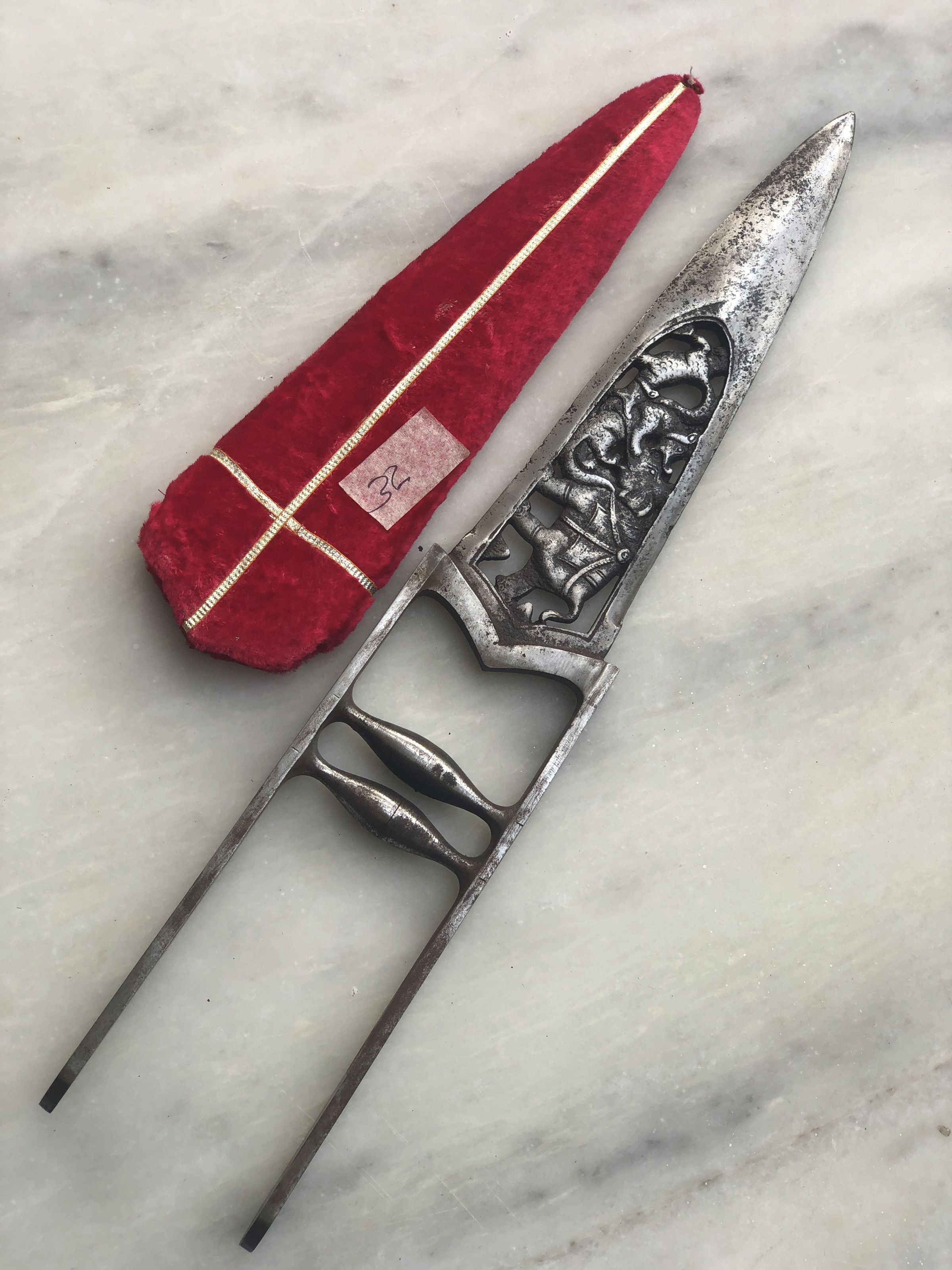 The image presents a ceremonial dagger alongside its red velvet sheath, both lying on a marble surface. The dagger features an elaborate handle with intricate silver filigree work and a pattern that suggests gothic or medieval influences. It has a metallic blade that tapers to a sharp point, and the grip design indicates that it might be a piece rooted in historical or ceremonial significance. The sheath is adorned with a simple, elegant cross pattern stitched in golden thread, adding a regal touch to the ensemble. A small, white paper tag marked with the number "32" in red ink is attached to the sheath, hinting that this item could be part of a collection or exhibit. From its remarkable craftsmanship, this dagger likely serves more as a decorative or collectible relic rather than a utilitarian tool, embodying artistic as well as historical values.