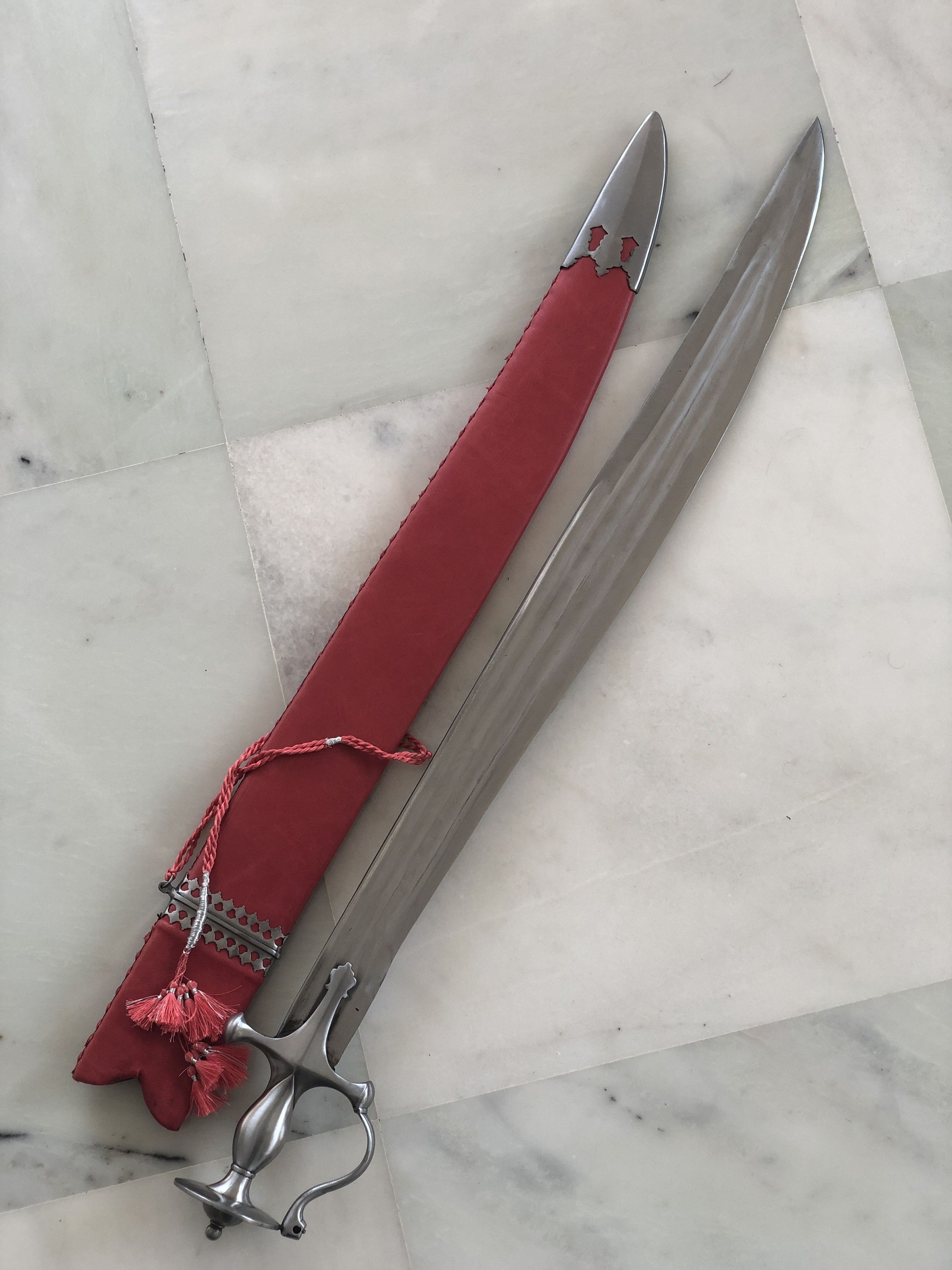 The image depicts a long, slender ceremonial sword lying on a marble floor. The sword's polished blade gleams with a mirrored finish, reflecting light and showing no signs of tarnish. It features an ornate handguard that is artistically crafted in a silver tone, with curves and openings that suggest a design both protective and decorative. This handguard connects to an elegant, possibly ergonomic handle, also metallic, hinting at a solid grip. Accompanying the sword is a scabbard enveloped in rich red fabric that appears slightly worn at its opening, giving a hint of frequent use or historical value. A decorative tassel with a braided rope accentuates the scabbard, adding a touch of ceremonial grandeur. This setup connotes a mix of functionality and ceremonial significance, possibly denoting the sword's use in historical or ritual contexts, suggesting not just a weapon, but a symbol of power and status. The marble backdrop provides a clean, stark contrast to the vivid red and