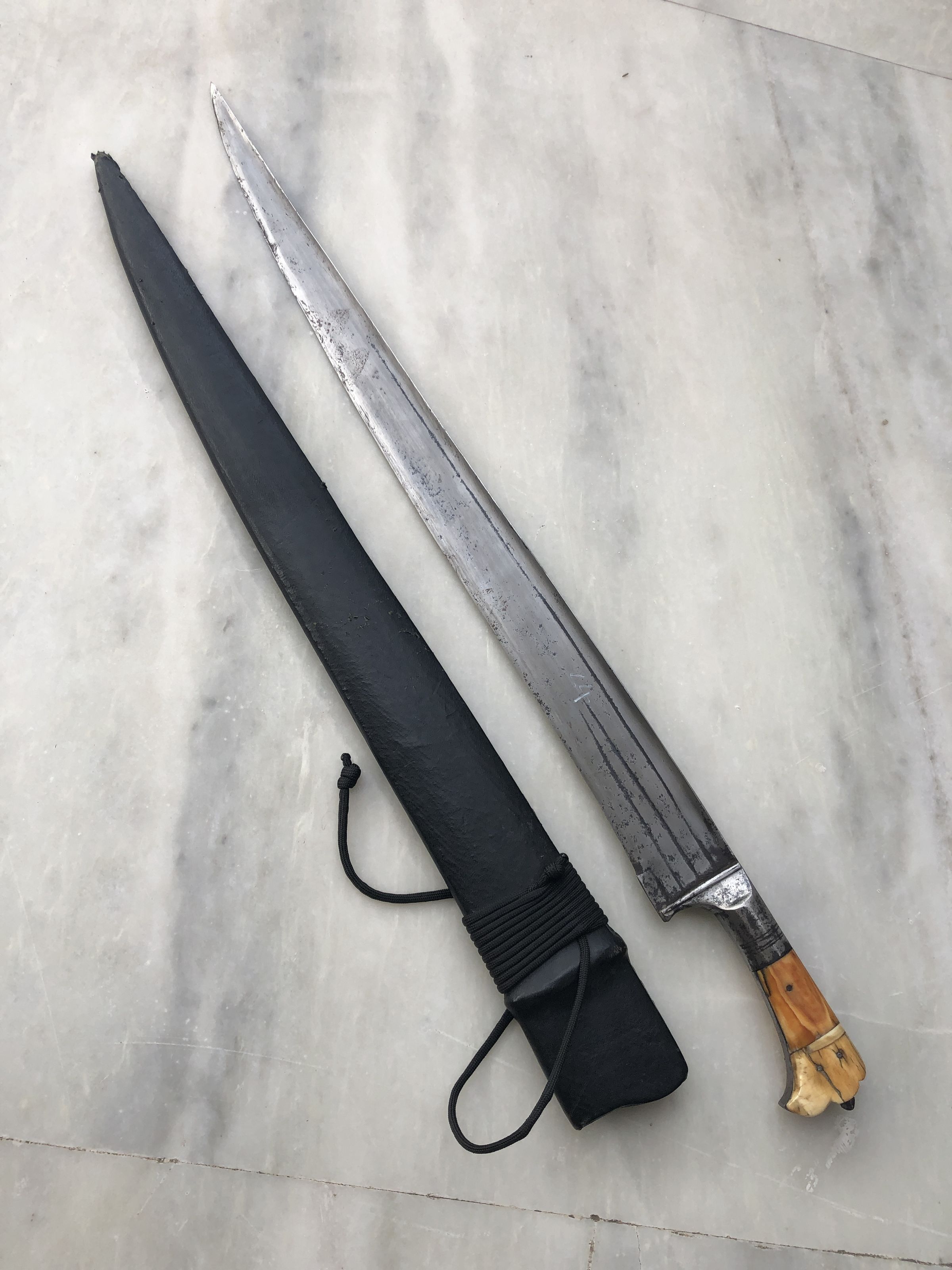 The image shows an Afghan Khyber knife with its blade and sheath displayed on a marble surface. The knife features a long, narrow, and slightly curved steel blade with visible signs of wear, which highlights its antiqued appearance. Attached to the blade is a sturdy handle crafted from a light-colored material, possibly horn or bone, fitted with metal accents that add a decorative touch. The handle's design ensures a good grip. The accompanying black sheath, crafted from leather, covers the entire length of the blade and is secured with a black cord wound tightly around it, adding practicality and a traditional look to the ensemble. This piece reflects a mix of utilitarian function and intricate craftsmanship, characteristic of such historical weapons from the region.