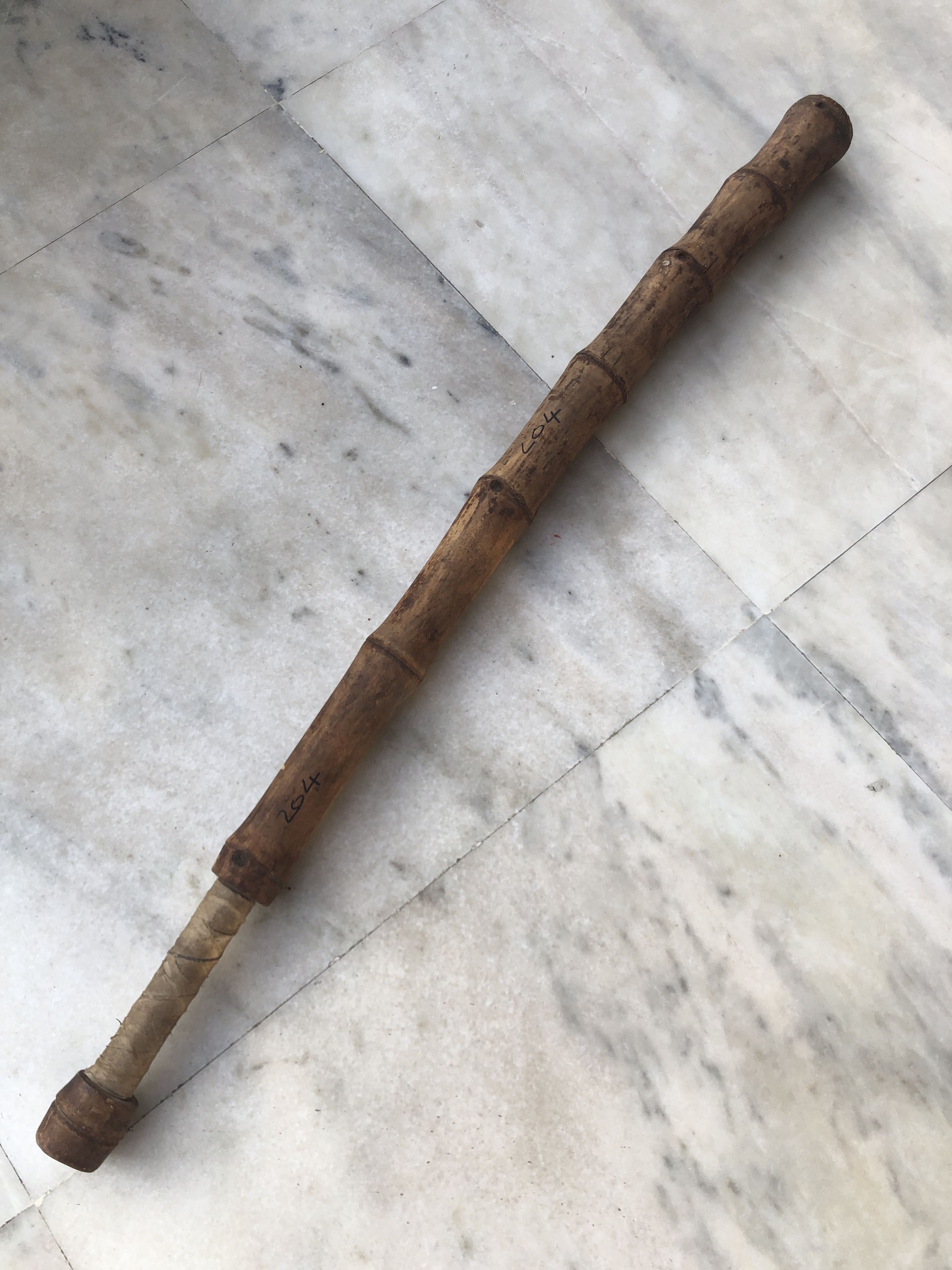 The image depicts an elongated wooden object, resembling a traditional staff or a walking stick, resting on a marbled floor that shows signs of wear and subtle stains. The staff appears aged, with a rich patina and distinctive graining evident along its surface, suggestive of considerable use over time. Sections of the staff are wrapped with a lighter-colored material, possibly leather or a similar binding, which could be intended to offer a better grip or to reinforce areas of frequent handling. Notably, the wood is marked with inscriptions or symbols near the center, possibly indicating ownership, significance, or markings from a particular craftsman or culture. The object's simplicity belies its possible historical and functional value, potentially serving as a tool, a ceremonial item, or a personal artifact from a bygone era.