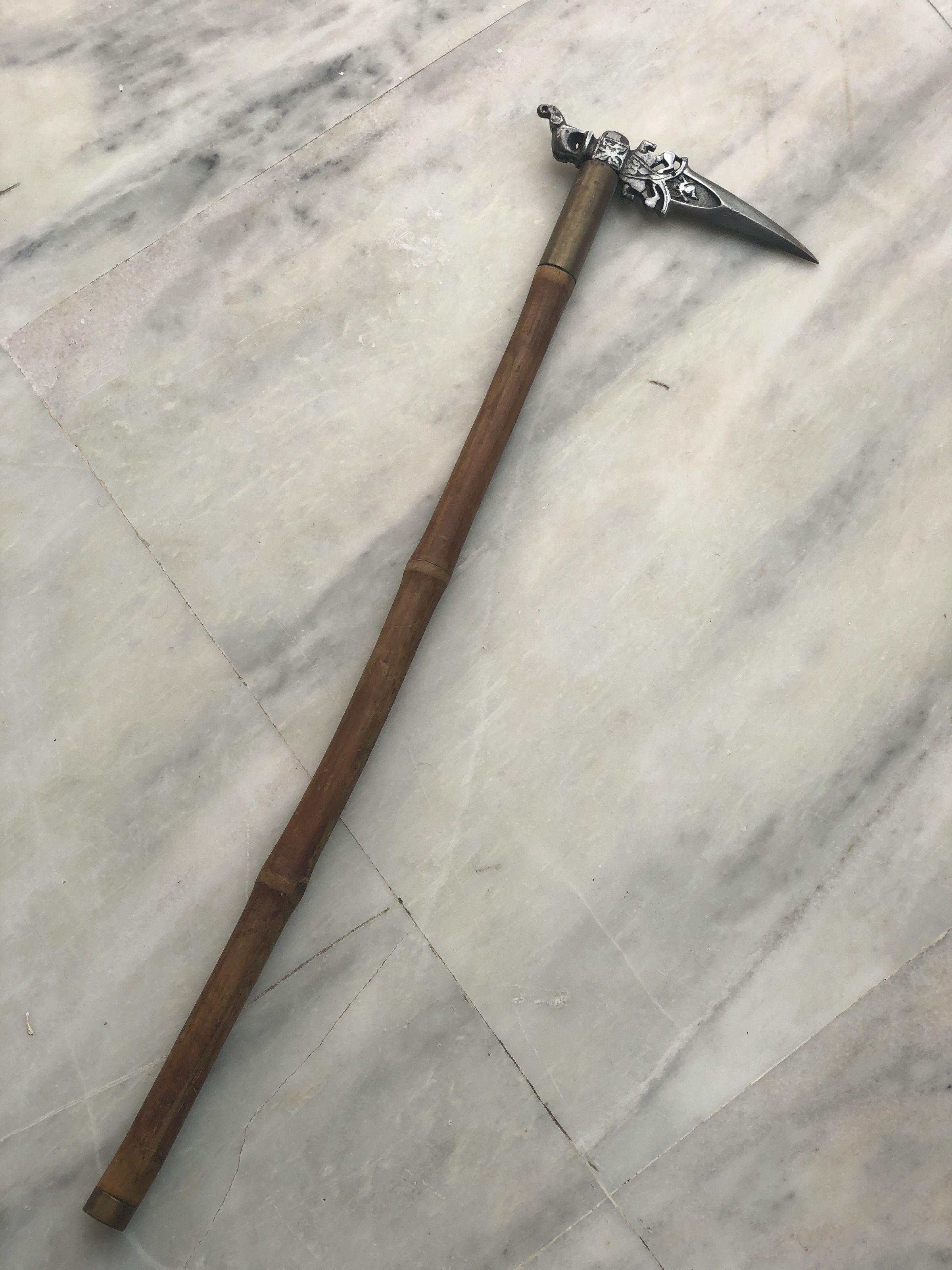 The image showcases a medieval pole weapon known as a Bec de Corbin or a Zagnol, lying on a marbled floor. The weapon features a long, smooth wooden shaft with a darker band wrapping around its midpoint, enhancing the grip. At one end, it is equipped with a striking head made from metal, designed intricately with ornamental details. The head includes a pointed beak-shaped protrusion suitable for piercing armor, and a flat hammer-like opposite side, intended for bludgeoning. This combination of features made the weapon versatile in historical combat. The craftsmanship reflects both the utilitarian and artistic aspects typical of medieval weaponry.