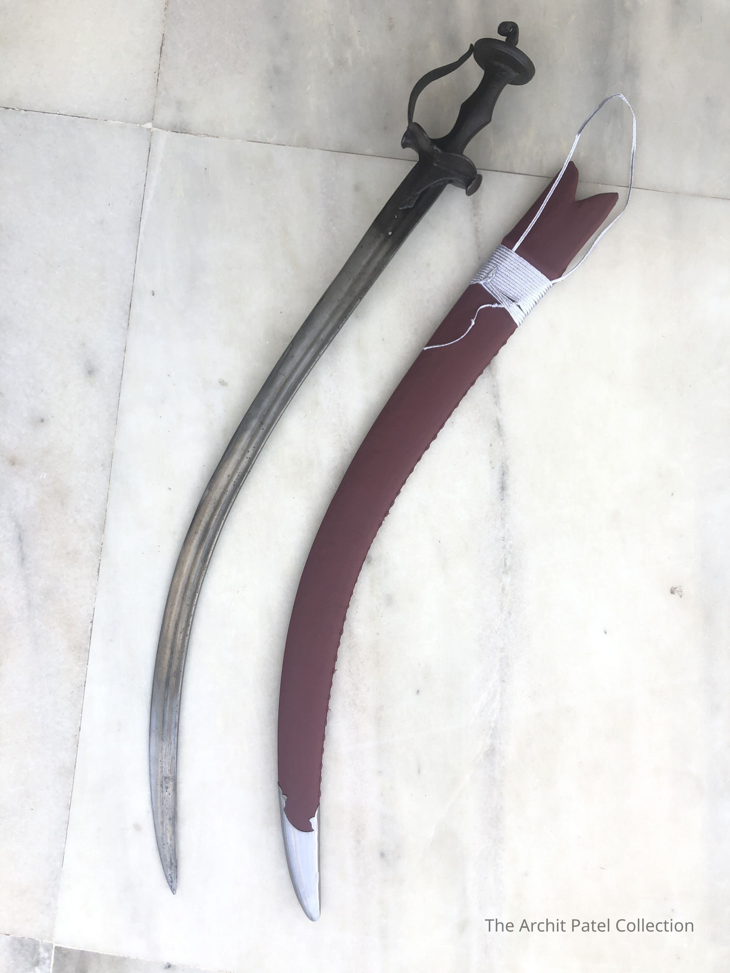 The image displays traditional sword with curved blades, laid on a white marble floor. On the left is a sword with a plain but elegant guard and a steel blade, possessing a slight curvature typical of sabers used in cavalry or military scenarios from past centuries. Its hilt is encased in a dark material, which might be wood or leather, providing a firm grip. The sheath on the right features a dark maroon scabbard, possibly made of leather or a synthetic material, designed to protect the blade and enhance safety when not in use. This sword's handle includes a protective guard that curves towards the blade, typical of designs intended to protect the hand during combat. Both pieces reflect historical weapon designs, likely replicas or antiques, and evoke a sense of historical military tradition and craftsmanship. They are oriented in parallel, suggesting a deliberate placement for display or examination.