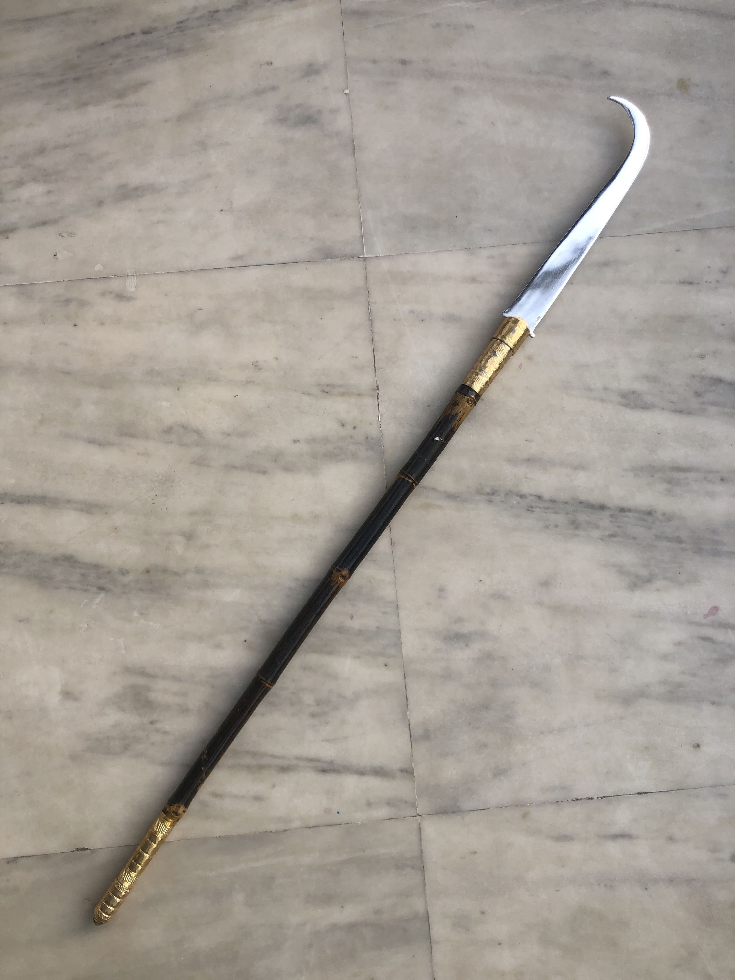 The image captures a traditional weapon with a striking curvaceous blade, set against a cool grey marble floor, hinting at both elegance and vintage aesthetics. At first glance, the prominent feature of the weapon is the cleanly polished, sharply curved blade that exudes a menacing yet controlled vibe, likely designed for fluid slicing motions given its pronounced arc. Moving towards the hilt, ornate details come into play, as segments of golden embellishments break the continuity of the otherwise dark lacquered handle. These gold areas add a regal touch reminiscent of historical weaponry, possibly indicating the weapon’s importance or ceremonial use. The laydown placement of the weapon on the marble brings an inherent contrast between the crafted metal and organic stone, pairing man-made artistry with the quiet background of nature’s own canvas. Each aspect of the weapon, from its tapered, sharp point to its luxuriously patterned handle, stresses a blend of functional design and aesthetic celebration—a piece that’s much more than mere armament
