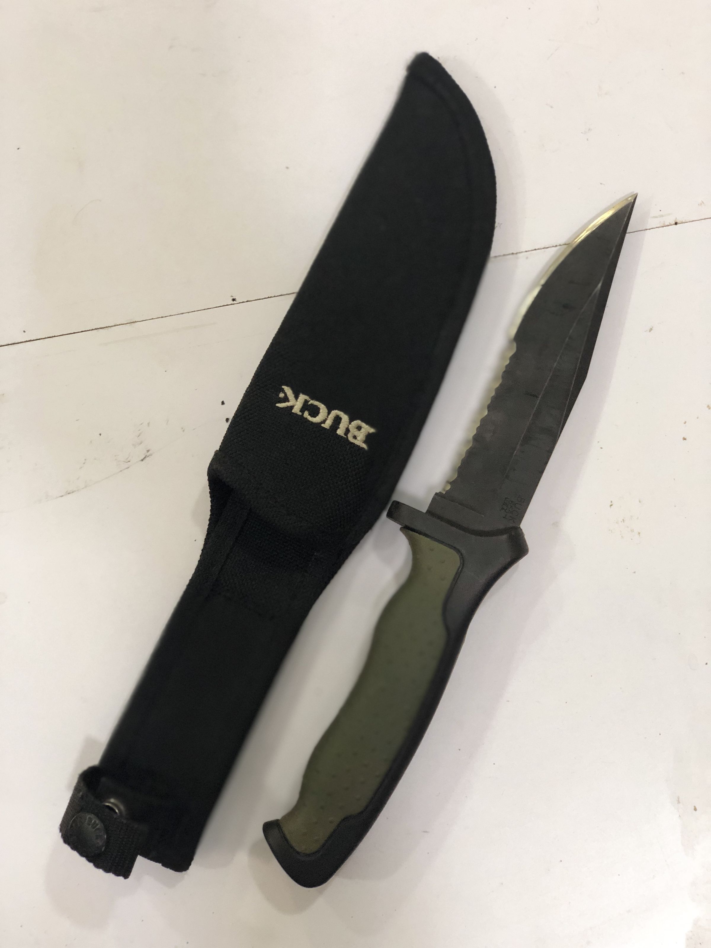 The image showcases a black tactical knife partially withdrawn from its black, fabric sheath which is emblazoned with the word "BUCK" in bold, white letters. The knife features a rugged design with a serrated blade near the handle, which is crafted with a curve to enhance grip. The handle itself is green with black outlines, ergonomically shaped with indentations to fit fingers comfortably, aiding in improved handling and safety. The sheath includes a loop for easy attachment to a belt, making it practical for outdoor activities. The floor beneath the knife appears to be a well-worn, white surface with visible marks and scratches, suggesting frequent use or storage in this spot.