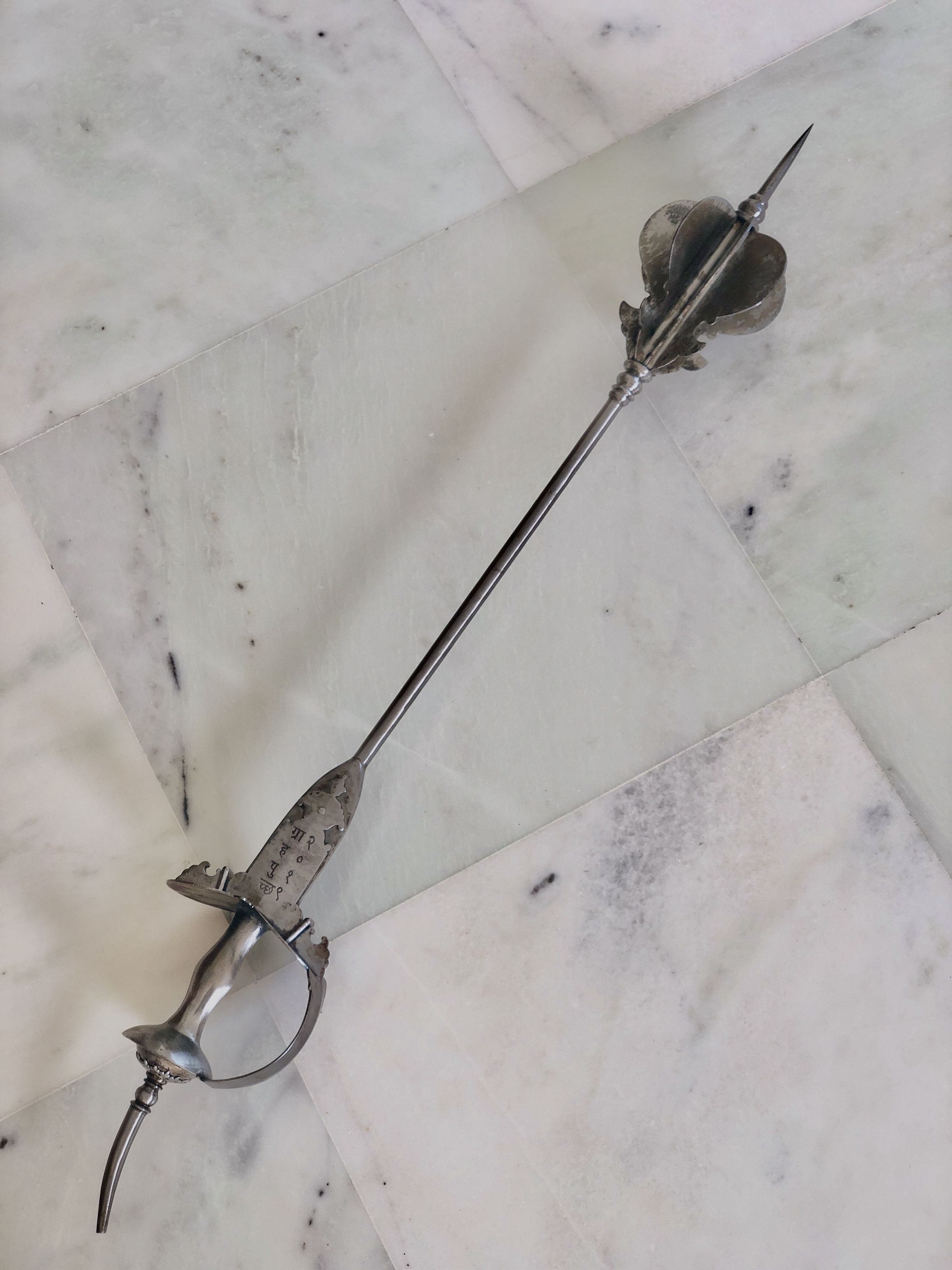 The image displays an elegant and intricately designed mace, resting diagonally across a marble floor with white and gray veining. This weapon features a detailed basket hilt that encloses almost the entire grip, providing substantial hand protection. The hilt is ornately crafted with metal that shows signs of wear and aging, suggesting it might be an antique or part of a historical collection. At the guard, there is an oval ring which further enhances the sword’s defensive capabilities. The shaft itself tapers to a sharp point, optimized for thrusting, which aligns with the characteristics of a rapier, commonly used in the Renaissance and later for dueling and as a status symbol. The overall appearance of the sword, combined with its placement on the sophisticated marble flooring, highlights its significance, possibly as a collectible item or a decorative piece meant to evoke a sense of historical valor and artistry.