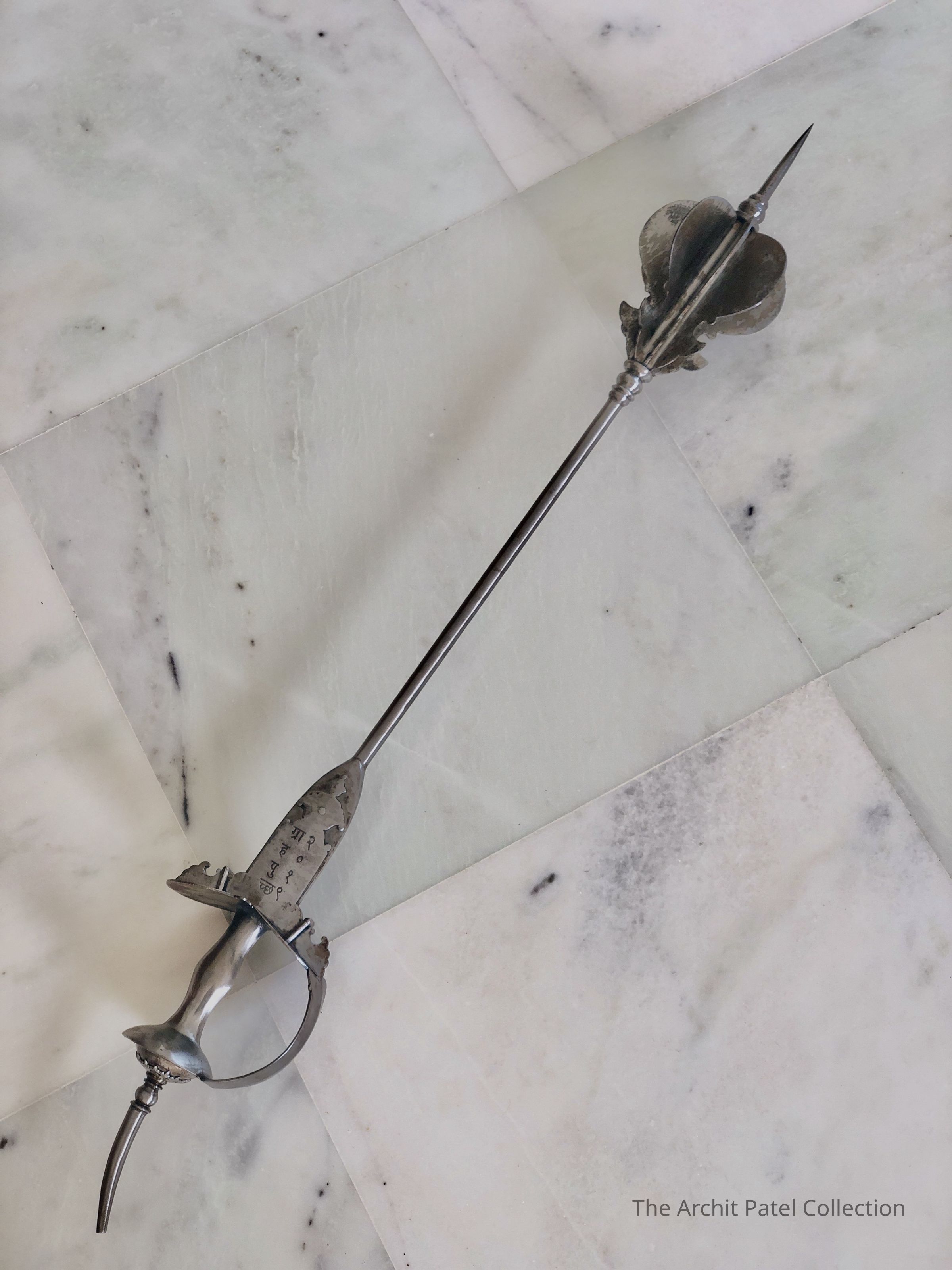The image displays an elegant and intricately designed mace, resting diagonally across a marble floor with white and gray veining. This weapon features a detailed basket hilt that encloses almost the entire grip, providing substantial hand protection. The hilt is ornately crafted with metal that shows signs of wear and aging, suggesting it might be an antique or part of a historical collection. At the guard, there is an oval ring which further enhances the sword’s defensive capabilities. The shaft itself tapers to a sharp point, optimized for thrusting, which aligns with the characteristics of a rapier, commonly used in the Renaissance and later for dueling and as a status symbol. The overall appearance of the sword, combined with its placement on the sophisticated marble flooring, highlights its significance, possibly as a collectible item or a decorative piece meant to evoke a sense of historical valor and artistry.