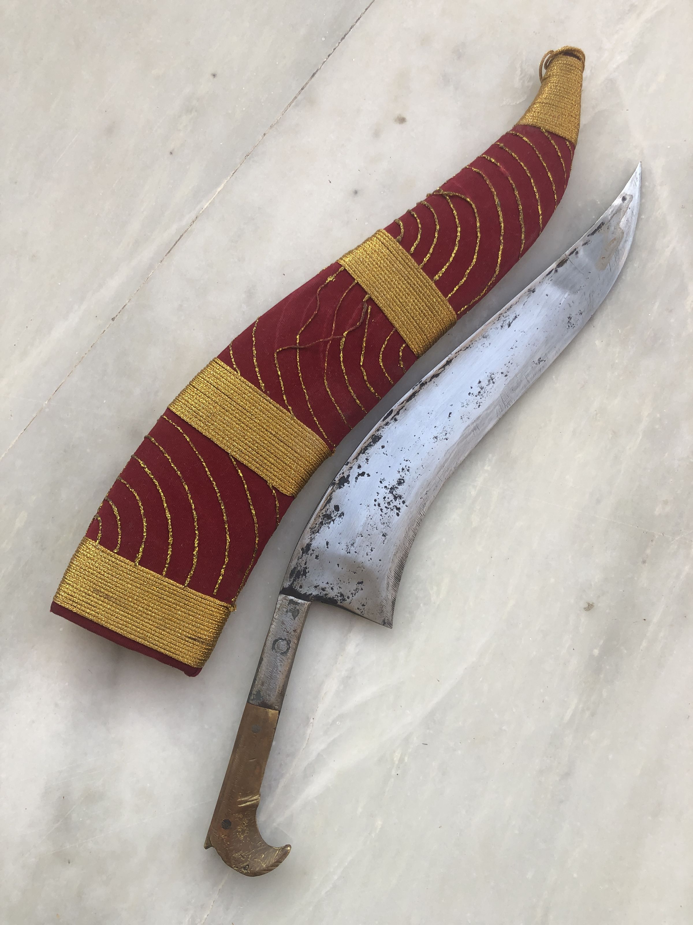 The image displays a traditional curved knife, often classified as a kukri or similar style blade, lying on a marble surface. This knife features a pronounced, elegantly curved steel blade with signs of wear and a patina indicative of age. The handle appears metallic, possibly brass, with a rounded, hooked end that enhances grip and control. Accompanying the blade is a striking sheath covered in a rich, burgundy fabric adorned with golden wave-like patterns, ensuring the blade is securely housed within. The juxtaposition of the ornate sheath and the rugged appearance of the blade suggests a blend of functionality and ceremonial use, characteristic of such historical artifacts.