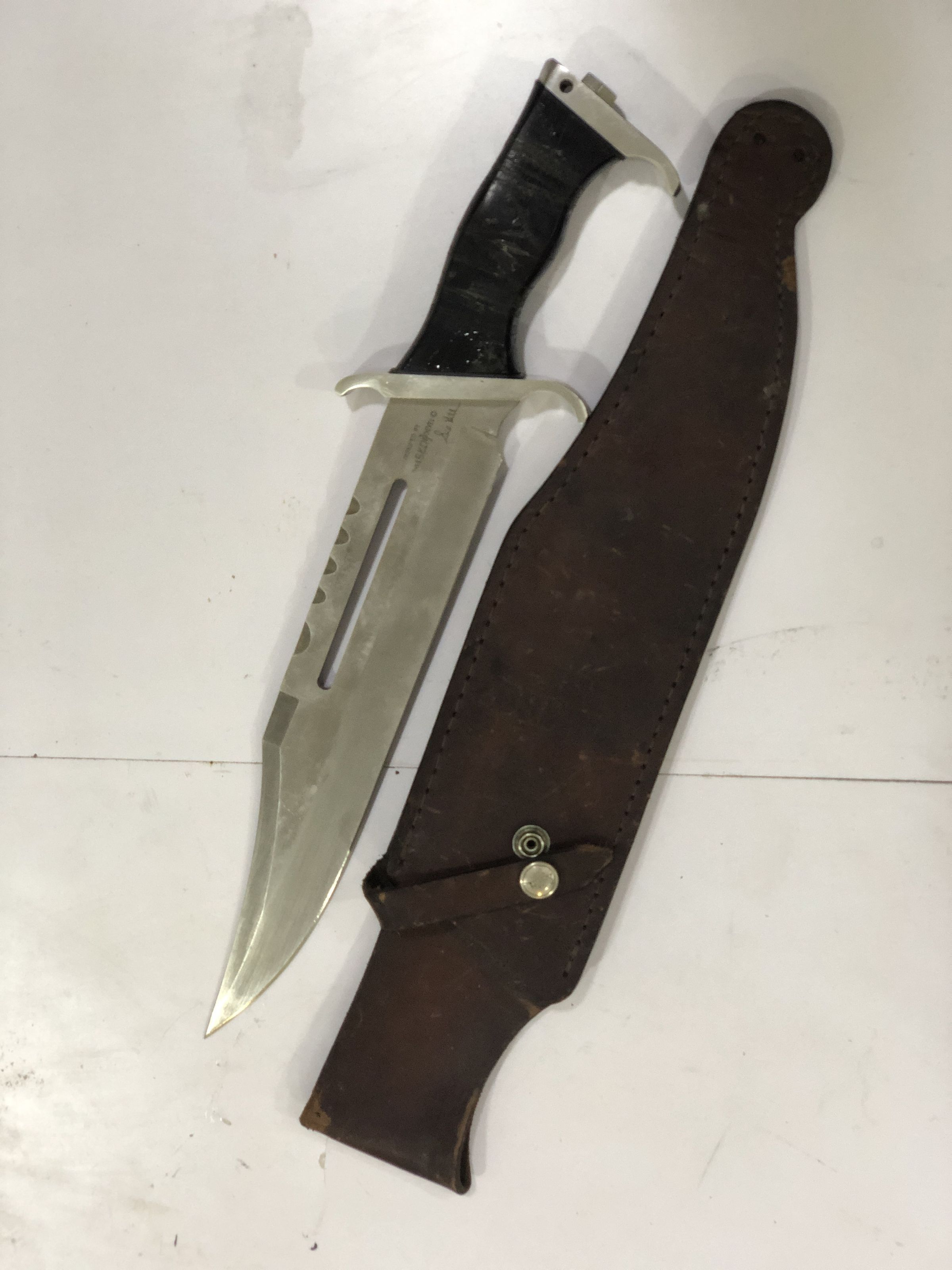 The image shows a large survival knife with a serrated blade and a black handle, lying next to its brown leather sheath on a white floor. The knife has a hole near the tip and a saw-like back edge, designed for multiple uses in survival situations. The sheath appears worn and features a metal snap button to secure the knife in place. The blade is marked with text near the handle, possibly indicating the maker or model of the knife. The overall condition and design suggest the knife is made for robust outdoor activities like camping or hunting.
