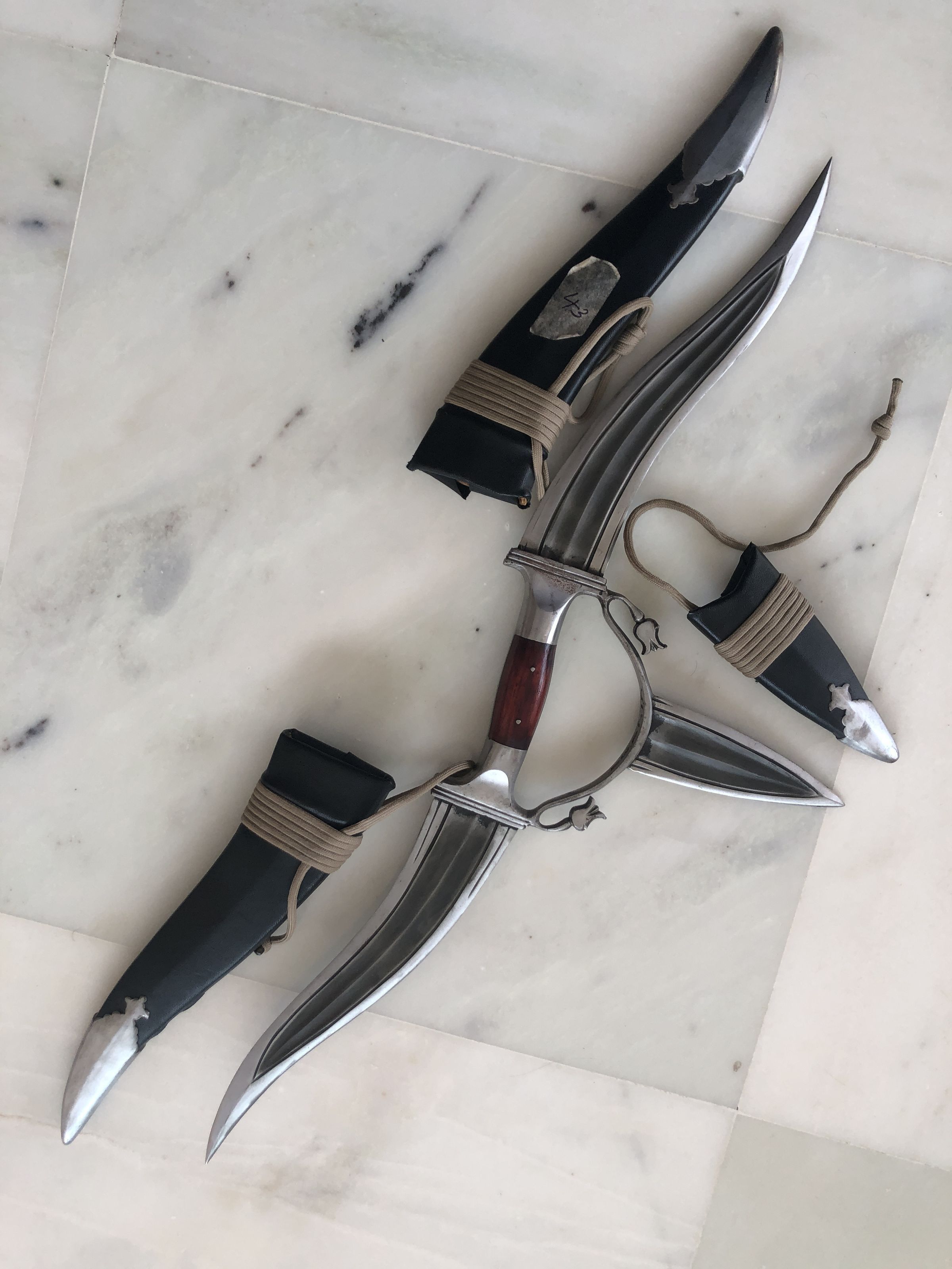 The image displays a collection of three ornate knives with unique and intricate designs, laid out on a marble floor. Two knives have curved blades and elaborately detailed handles, possibly signifying a ceremonial or decorative use, while the third knife is sheathed in a black cover with a simpler design, suggesting more practical usage. The craftsmanship indicates skilled metalwork and suggests cultural or historical significance. Each knife is complemented by distinctive features like embellished guards and handle details, enhancing their visual intrigue.