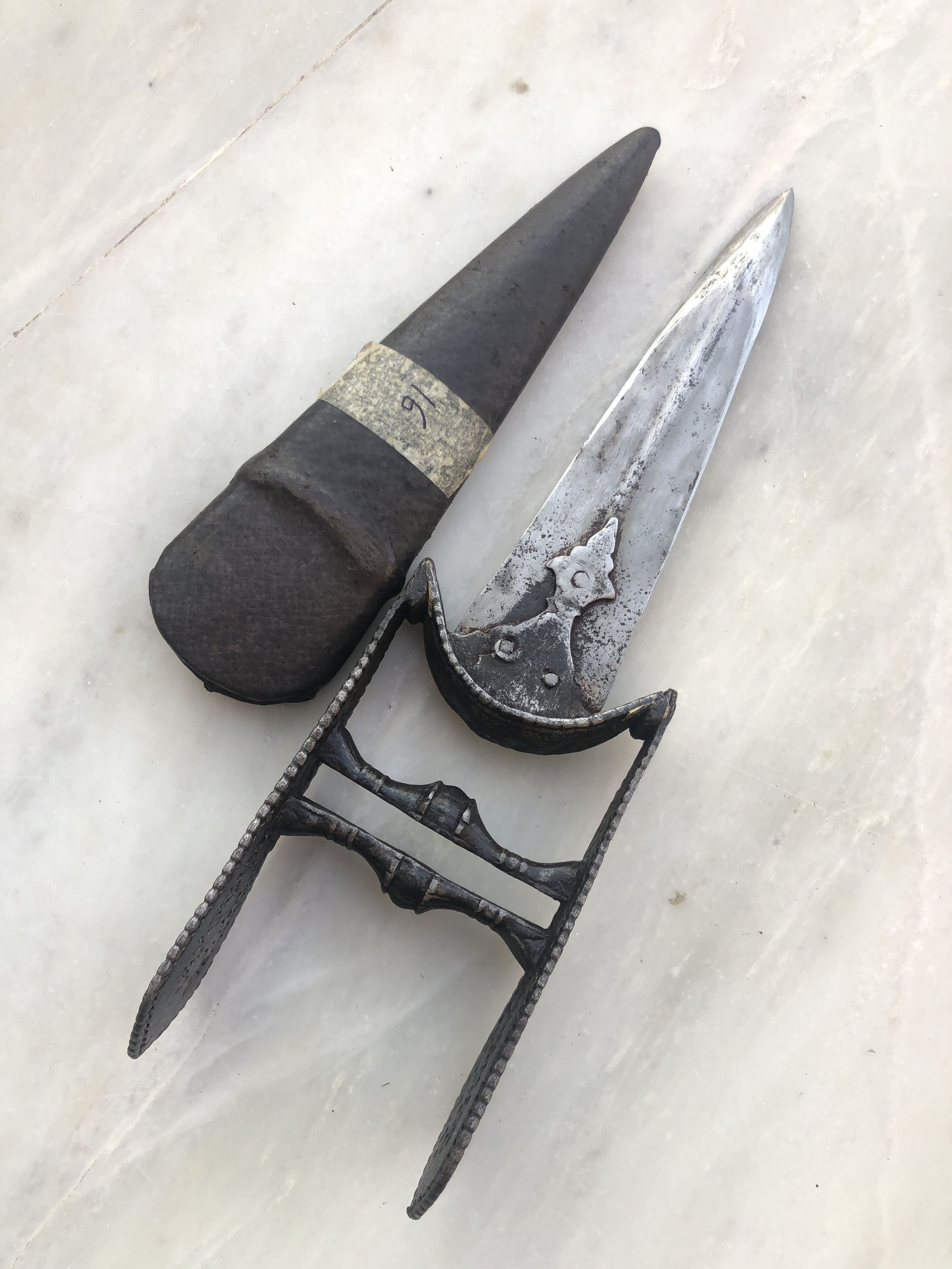 The image displays two antique-looking knives, each with distinct characteristics, laid on a light-colored marble surface. The sheath on the left features a simple, robust handle with a worn-out label marked with the number "69", attaching to a dark, unadorned blade. In contrast, the knife on the right flaunts a more intricate design, including an ornate handle with a detailed cross-guard and a blade adorned with a subtle floral pattern, demonstrating craftsmanship from perhaps a historical or ceremonial use.