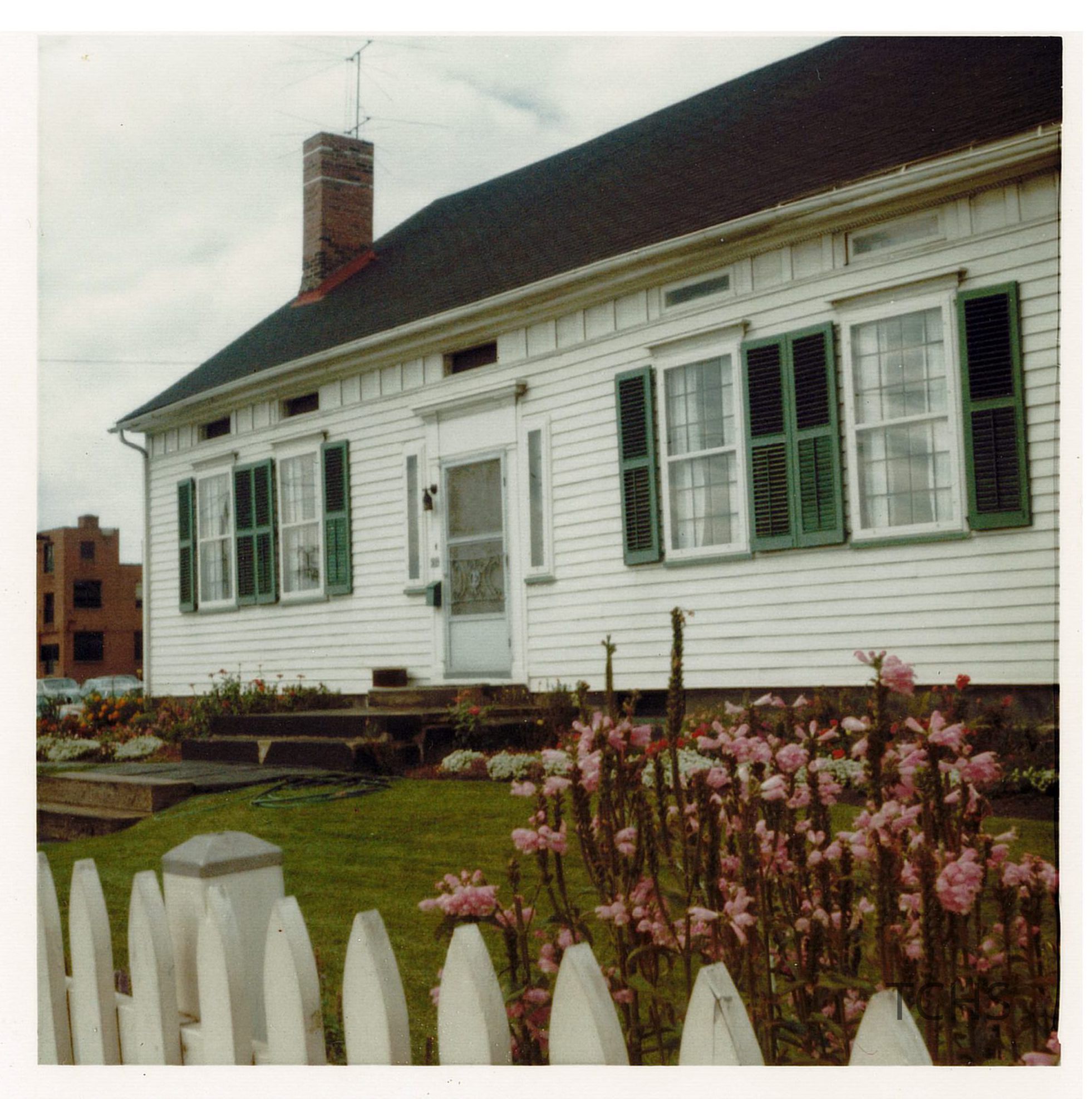 The image captures a charming, white clapboard house featuring a steeply pitched roof with dark shingles and a striking red brick chimney. It is adorned with green shutters, and the front door. A neatly maintained garden bursting with pink flowers lies before the house, partially enclosed by a crisp, white picket fence.