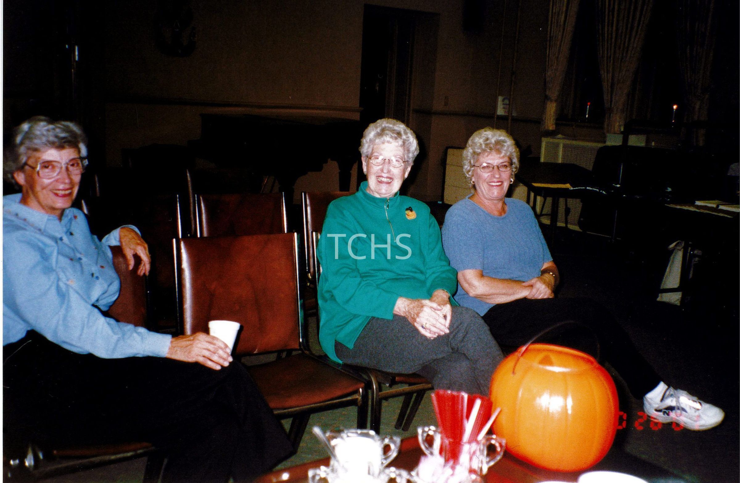 Three elderly women sit smiling in a warmly lit room, which appears set up for an event with brown folding chairs and a piano visible in the background. The lady on the left wears a light blue button-down shirt, the middle woman sports a green sweater with a decorative pin, and the one on the right is dressed in a casual blue top.