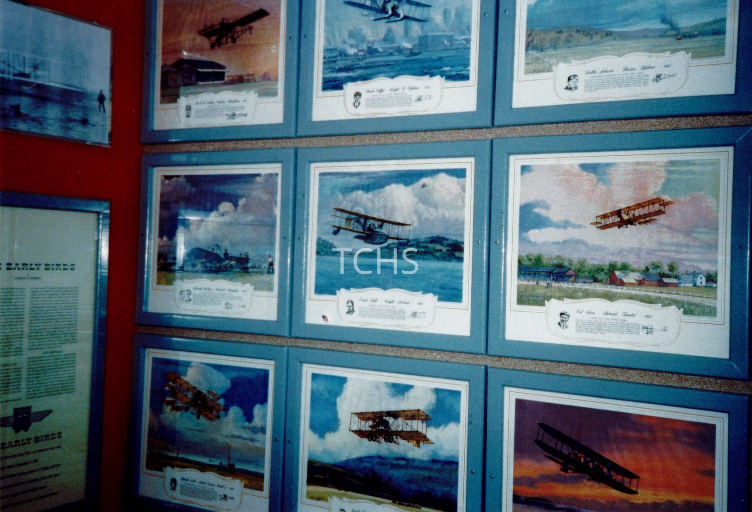 The image shows a collection of framed artworks displayed on a red wall, each depicting various historical aircraft in flight above different landscapes. Below each picture, a descriptive text plate seems to provide information about the aircraft and possibly the historical context, enhancing the educational value of the display. These paintings highlight the evolution and achievements in early aviation, capturing moments of airplanes over fields, water, and near buildings, portrayed against serene and moody skies.