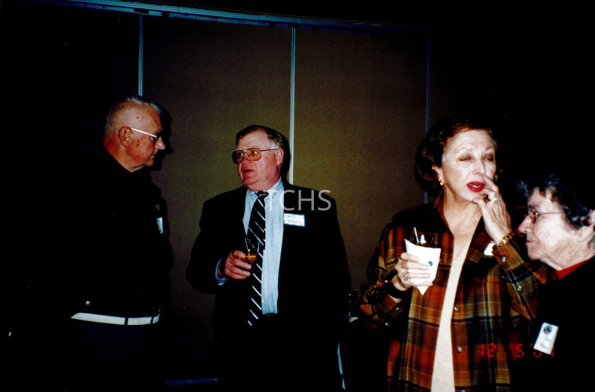 In the image, four elderly individuals are engaged in conversation at a social gathering inside a room with a basic, neutral interior. The men are dressed formally, one in a dark blazer and another sporting a striped tie, while the women are more casually attired, with one donning a vibrant plaid jacket and holding a drink. Their expressions and postures suggest a relaxed interaction during what appears to be a community or private event.
