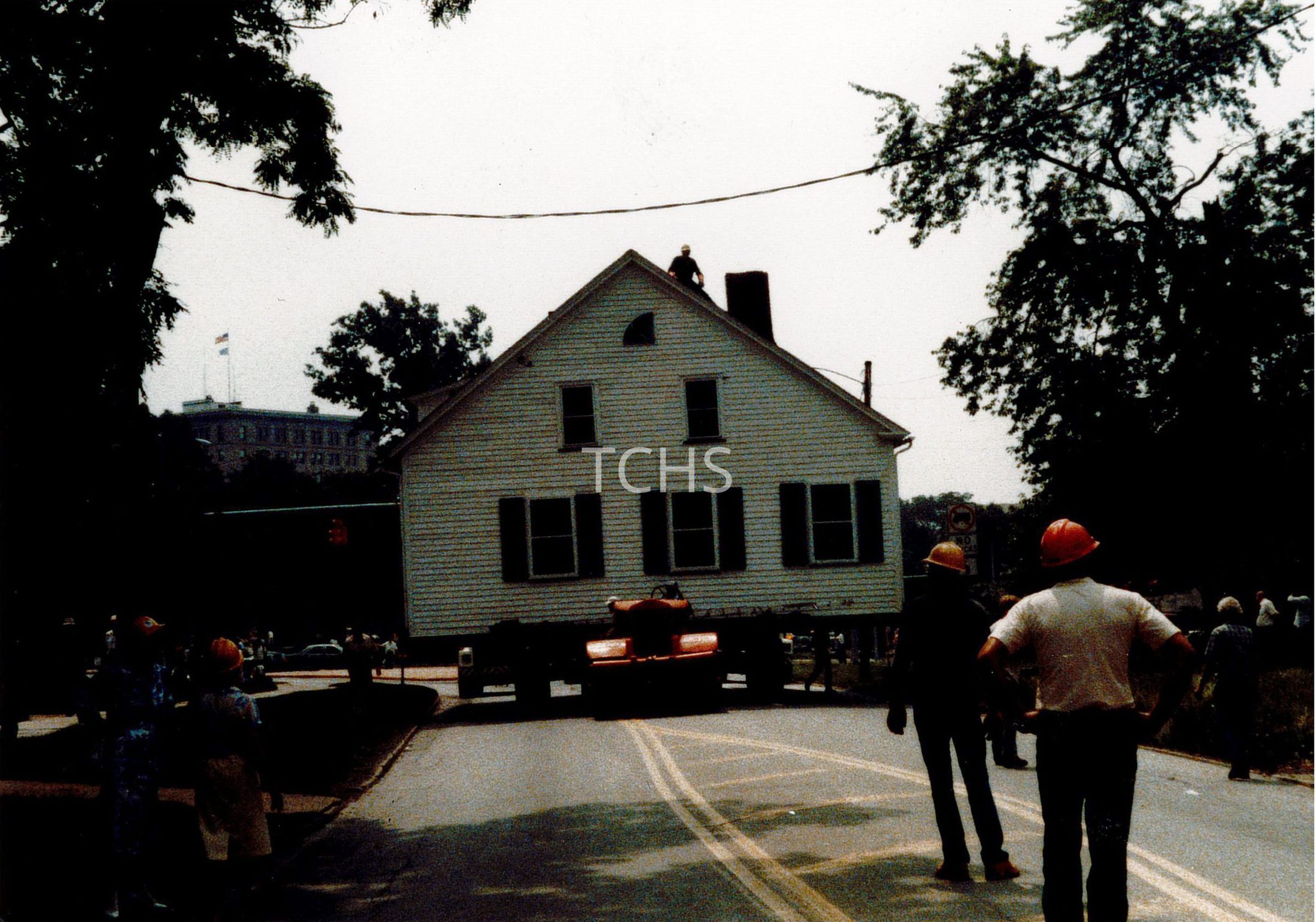 In the historical image, onlookers, some in hard hats and others casually dressed, gather around a large white house being transported across a road on a massive truck. The house, appearing quaint with its classic American architecture, contrasts starkly with the industrial nature of the heavy machinery utilized for its relocation. A person is visible on the rooftop, possibly overseeing the operation, while the tree-lined street and buildings in the background hint at a small, possibly rural town setting.