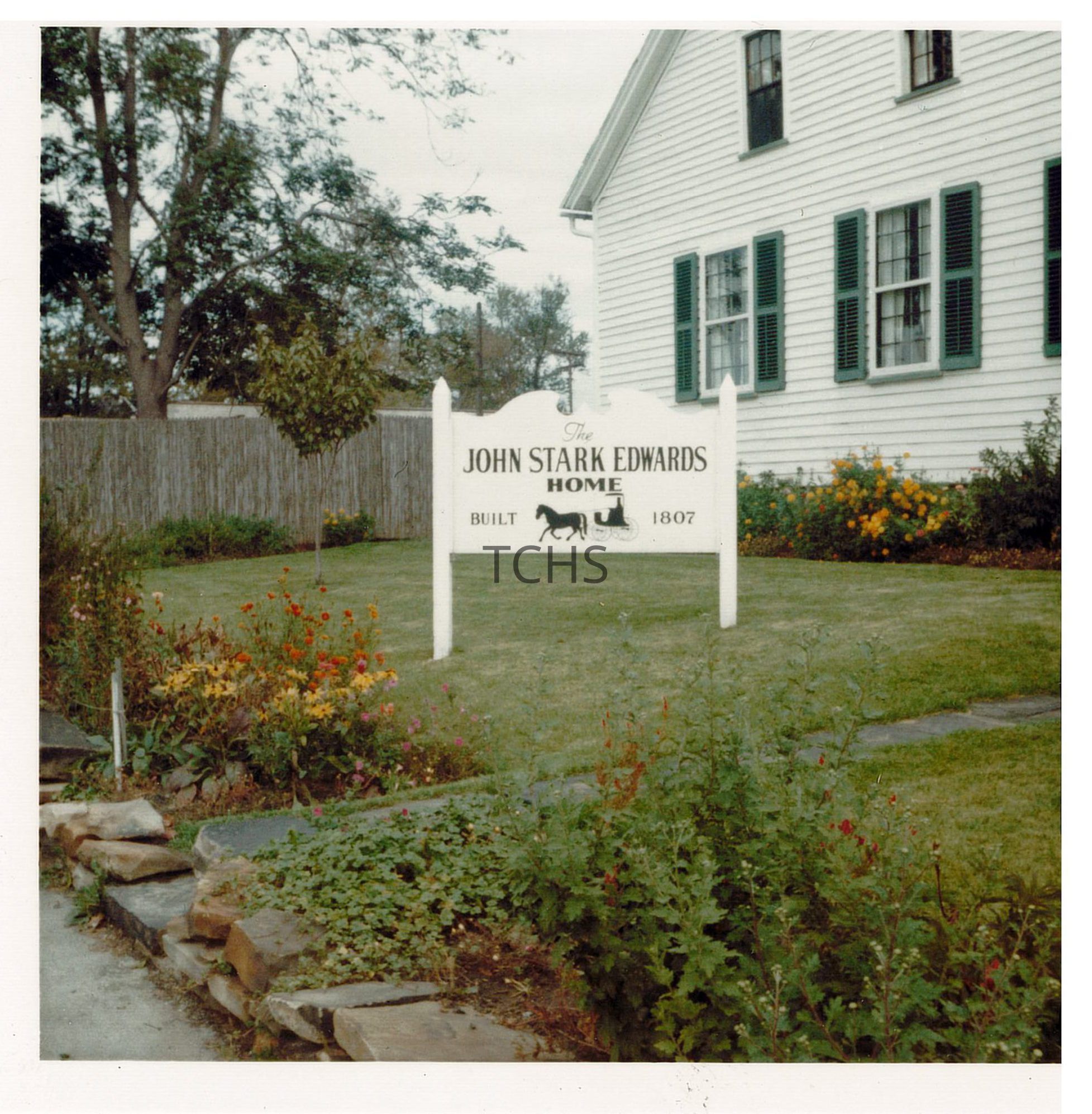 The image depicts the John Stark Edwards Home, constructed in 1807, featuring a charming white house with green shutters surrounded by a well-kept garden and a stone pathway. A white sign with black lettering stands prominently in the foreground, amidst lush grass and colorful flowers, providing historical information about the building. A wooden fence and trees can be seen in the background, enhancing the serene suburban setting.