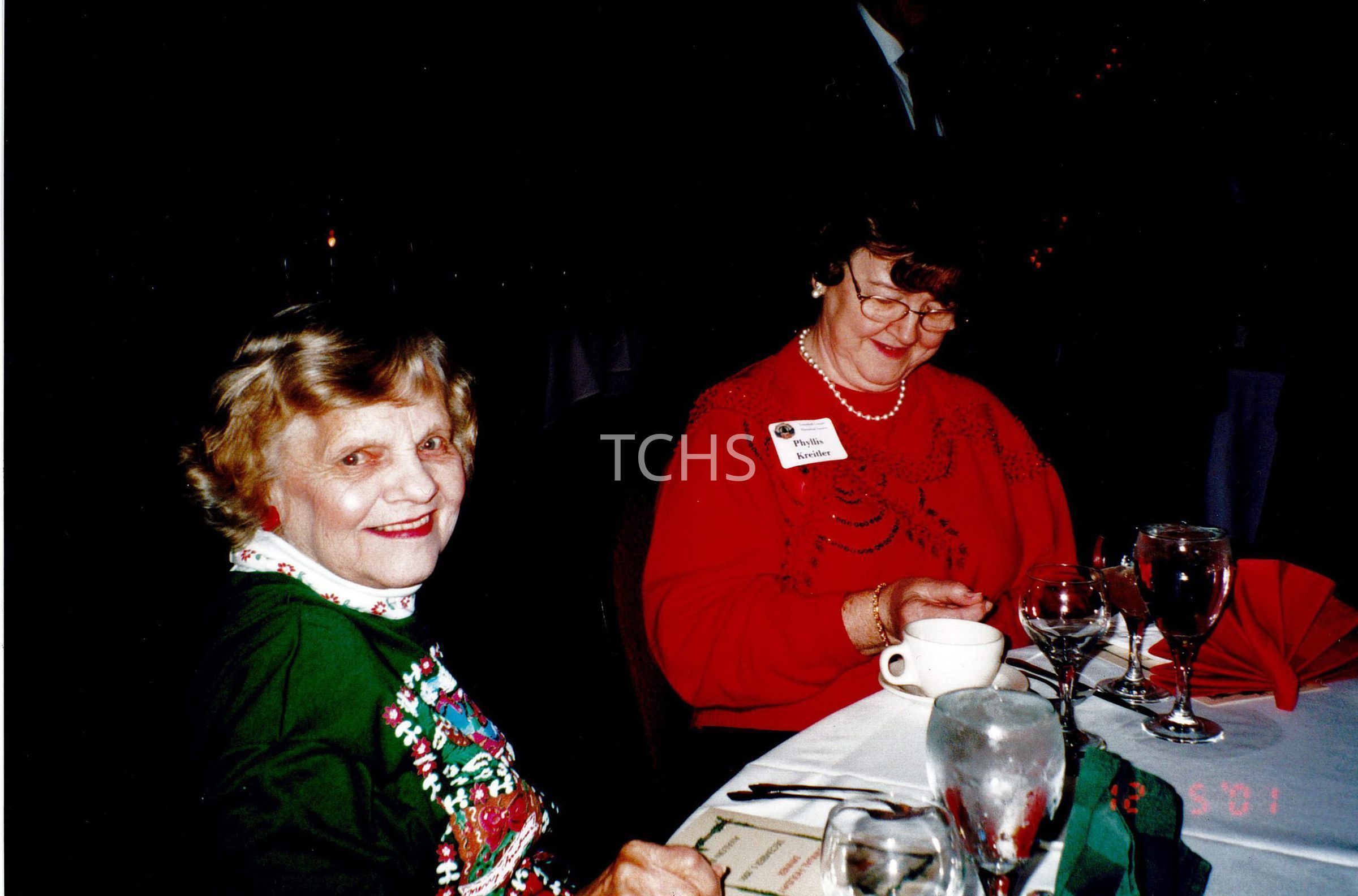 In the image, two women are smiling jovially during a festive dinner gathering, seated at a table adorned with empty wine glasses and a white tablecloth. One woman, clad in a red sweater with a pearl necklace, is glancing down, possibly reading a menu, while the other, wearing a festive green jacket with a colorful collar, looks directly at the camera, her face lit with a warm, inviting smile.