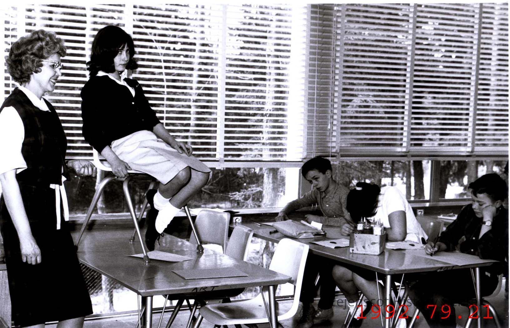 Sketching a Live Model: A black and white photograph of Mrs. Mary Ganske's art class at Brunswick Junior High School sketching a live model. Mrs. Ganske is seen standing to the left observing her students, while the live model sits on a chair placed on a table directly in front of three students engrossed in their sketches. Dated 1962.
