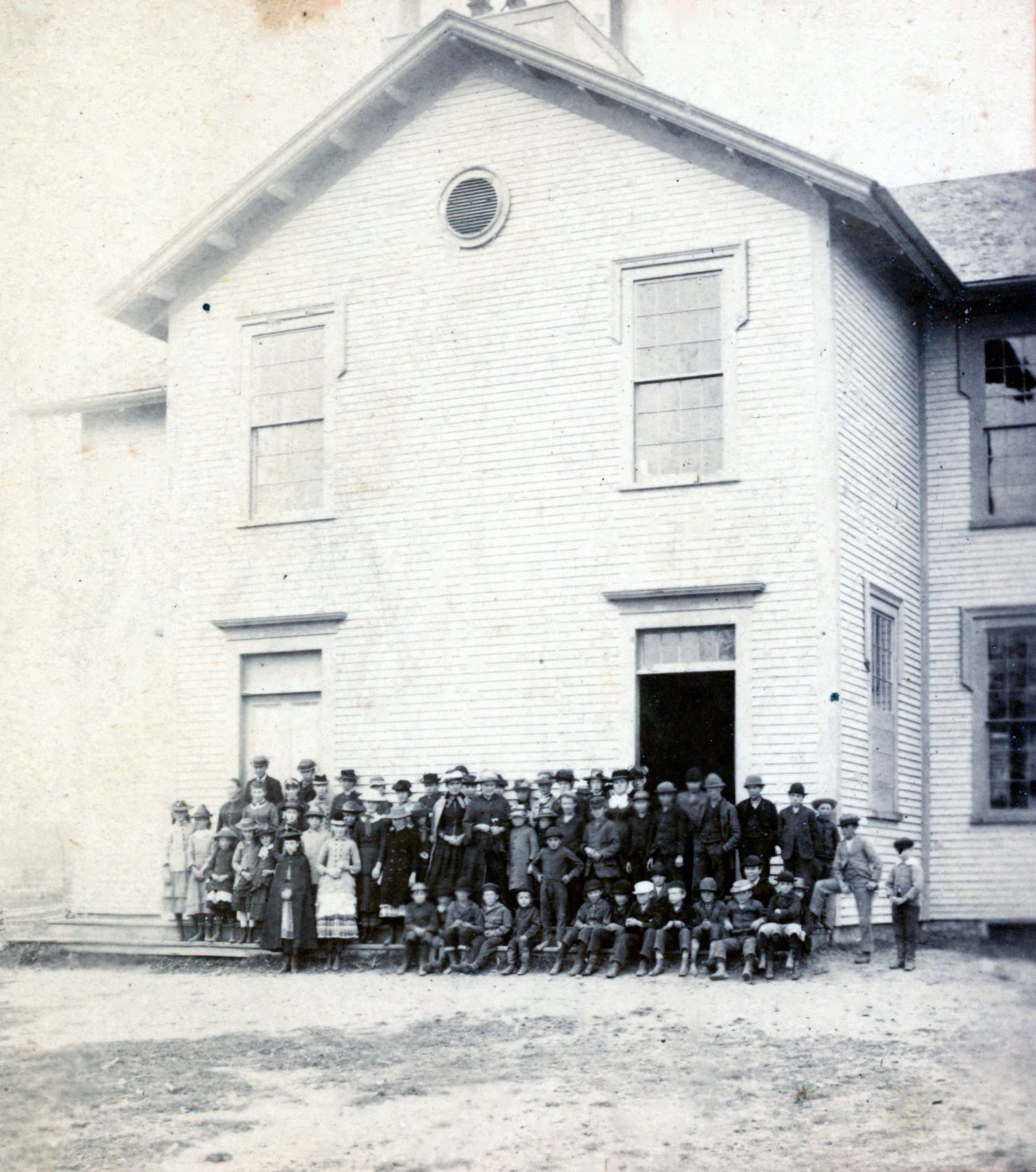 Students at the Dennysville Town School, c. 1910; Elementary and high school students gathered with their teachers on the front steps of the Dennysville Town School around 1910. The primary school students, in front, were taught in one downstairs classroom, while the high school students received instruction in the other. Judging from the caps and warm cloaks, the photograph must have been taken on a cold day. The school bell visible in the belfry above was relocated to the lawn of the Lincoln Memorial Library next door when the schoolhouse was demolished in 1964.
