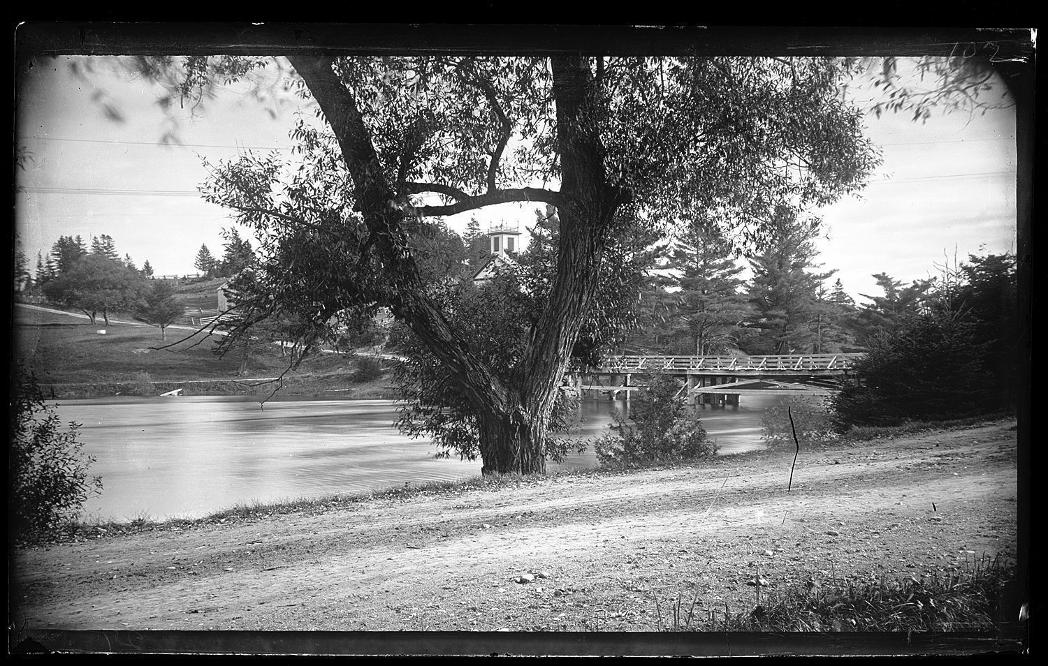 The Lower Bridge over the Dennys River in Summer; The Lower Bridge is seen from the road in front of Dennysville's Lincoln House with the Methodist Episcopal Church and other structures visible on the Edmunds side of the Dennys River, in this photograph by Dr. John P. Sheahan, c. 1880.