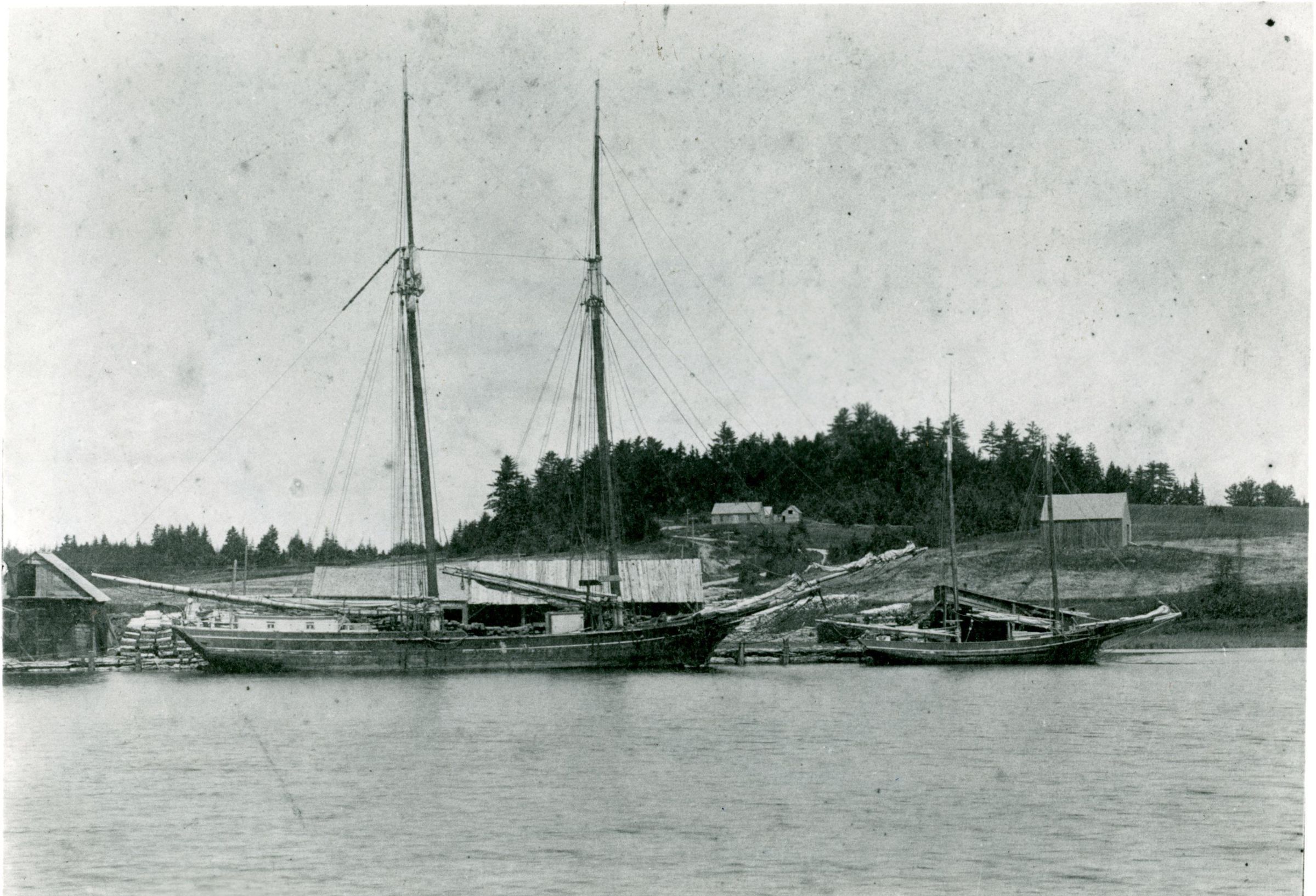 Schooner and packet at the Lincoln Wharf on the Dennys River, with Corn Hill in view.; Edmunds Lincoln's modest house and a barn are visible on Corn hill, behind the schooner and packet tied up at Lincoln's Wharf on the Dennys River in Dennysville, Maine, c. 1890.