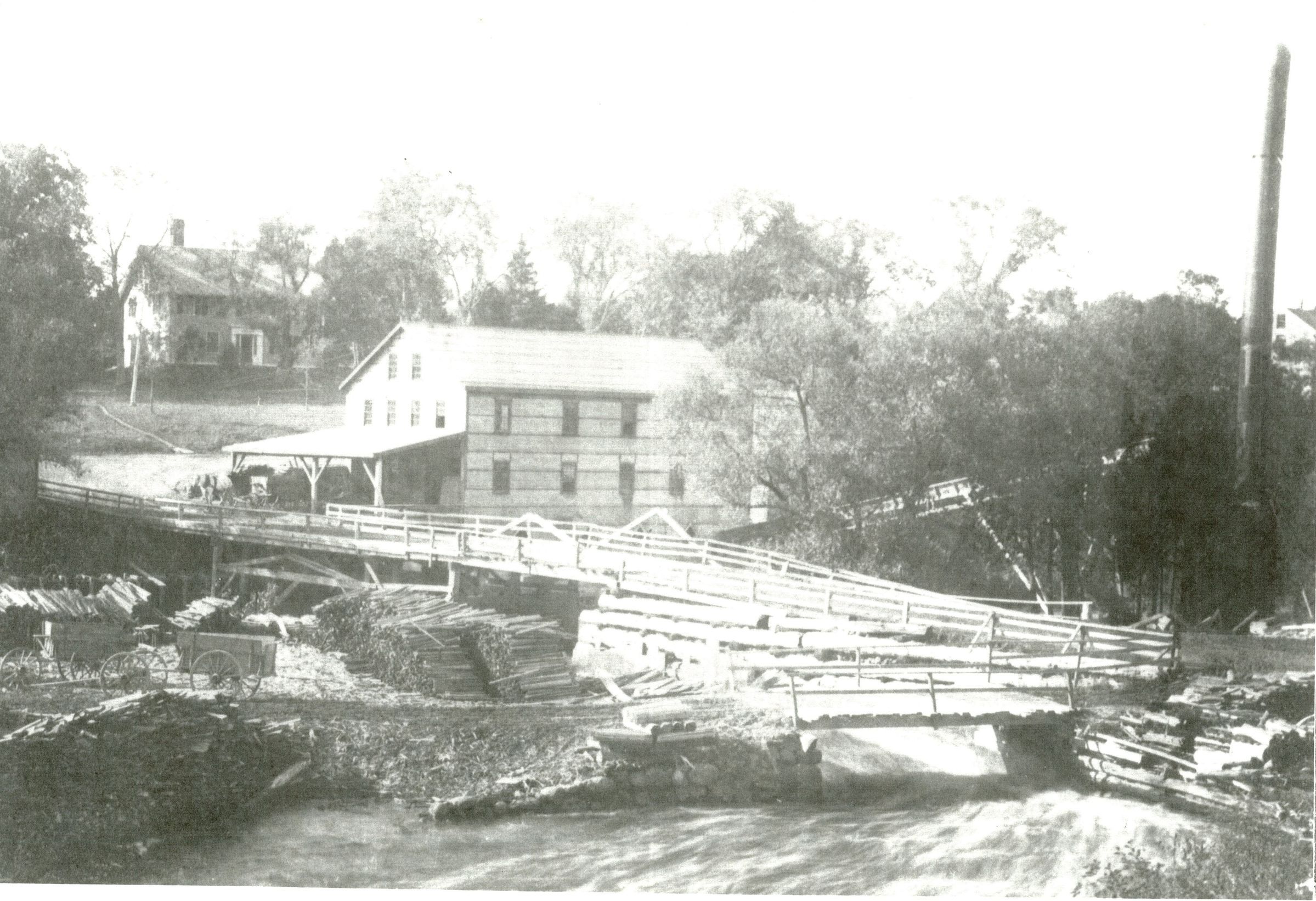 Dennysville Lumber Company in Dennysville, Maine; Water sluices around the mill dam, which powers the newly rebuilt lumber mill in 1907, with the Mill House. where the company managers lived on the hill beyond. A waste burner with conveyor belt stands on the Edmunds side of the river to consume the refuse of the mill.