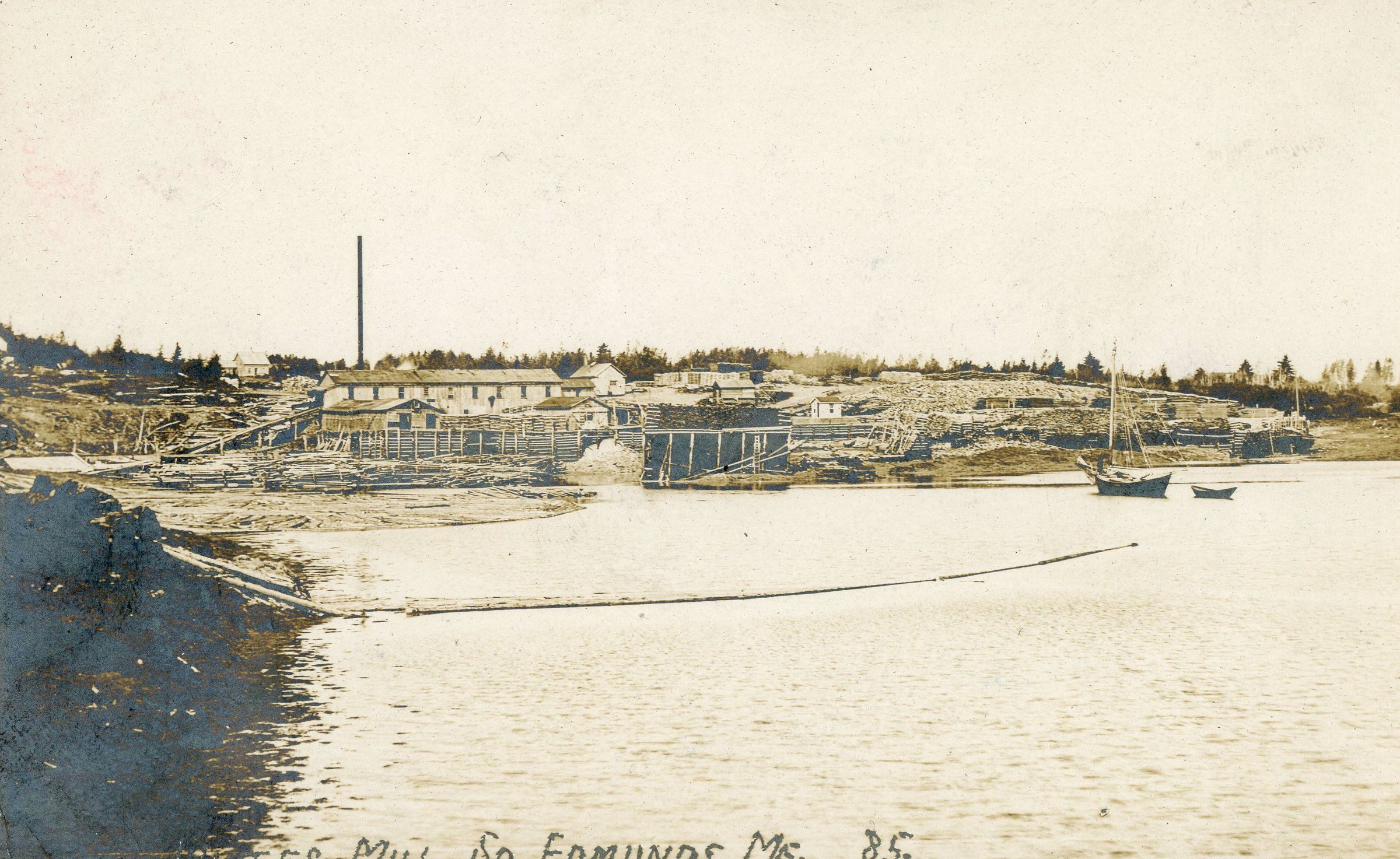E.I White's Steam Mill in Edmunds, Maine. c. 1905, viewed from Hallowell's Island; An boom is anchored to Hallowell's Island to retrieve any stray logs that may escape the timbers confined closer to the mill.